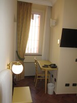 A small room with a single table, a chair, a lamp, a TV mounted on the wall, and a window with curtains.