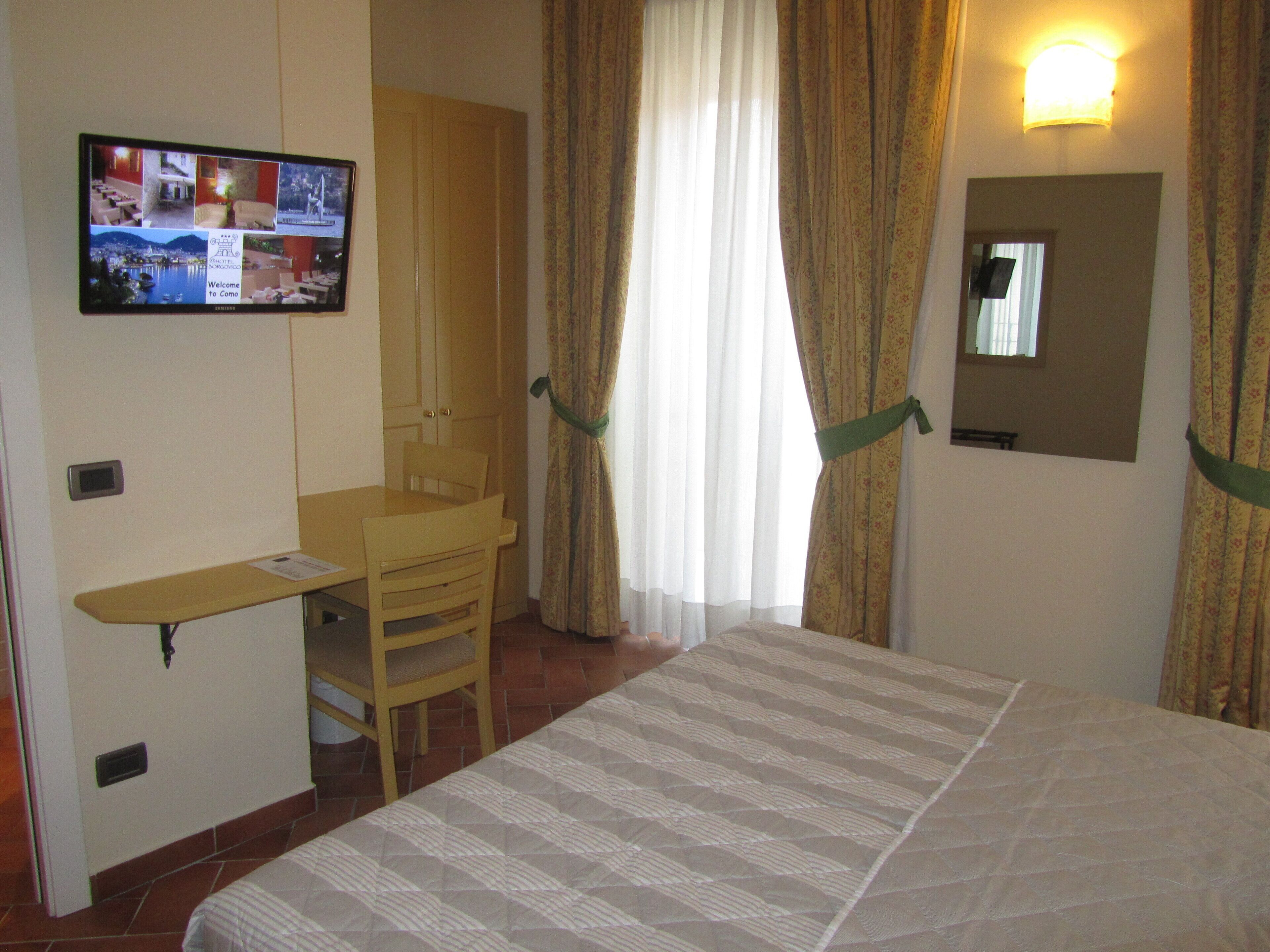 Habitación de hotel con cama, escritorio, silla, televisor mostrando un paisaje urbano y ventana con cortinas.