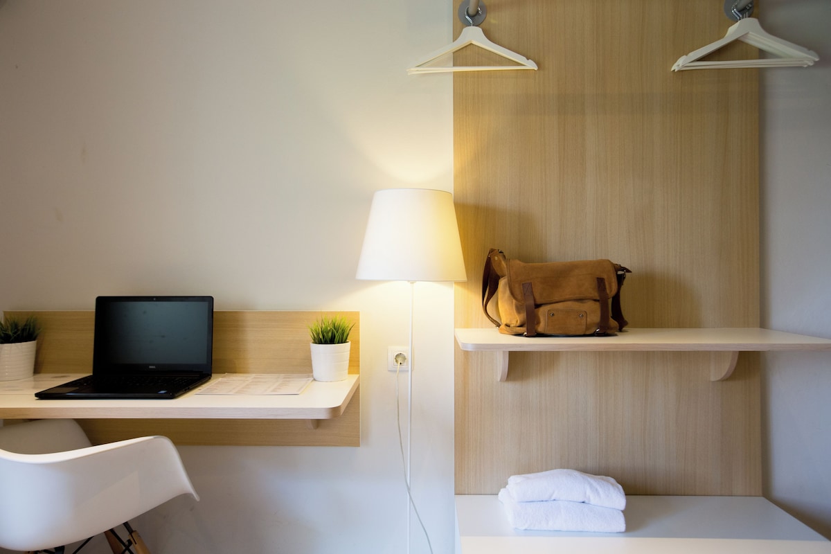 A hotel room with a desk, laptop, lamp, hanging clothes rack, and a small shelf with a bag and towels.
