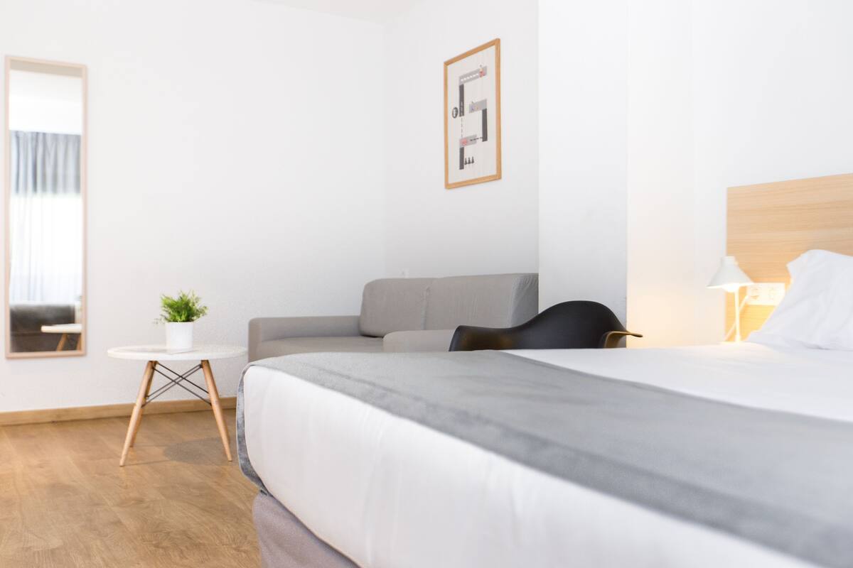 Une chambre d’hôtel moderne avec un grand lit, une petite table ronde agrémentée d’une plante, un canapé gris et un tableau encadré au mur.