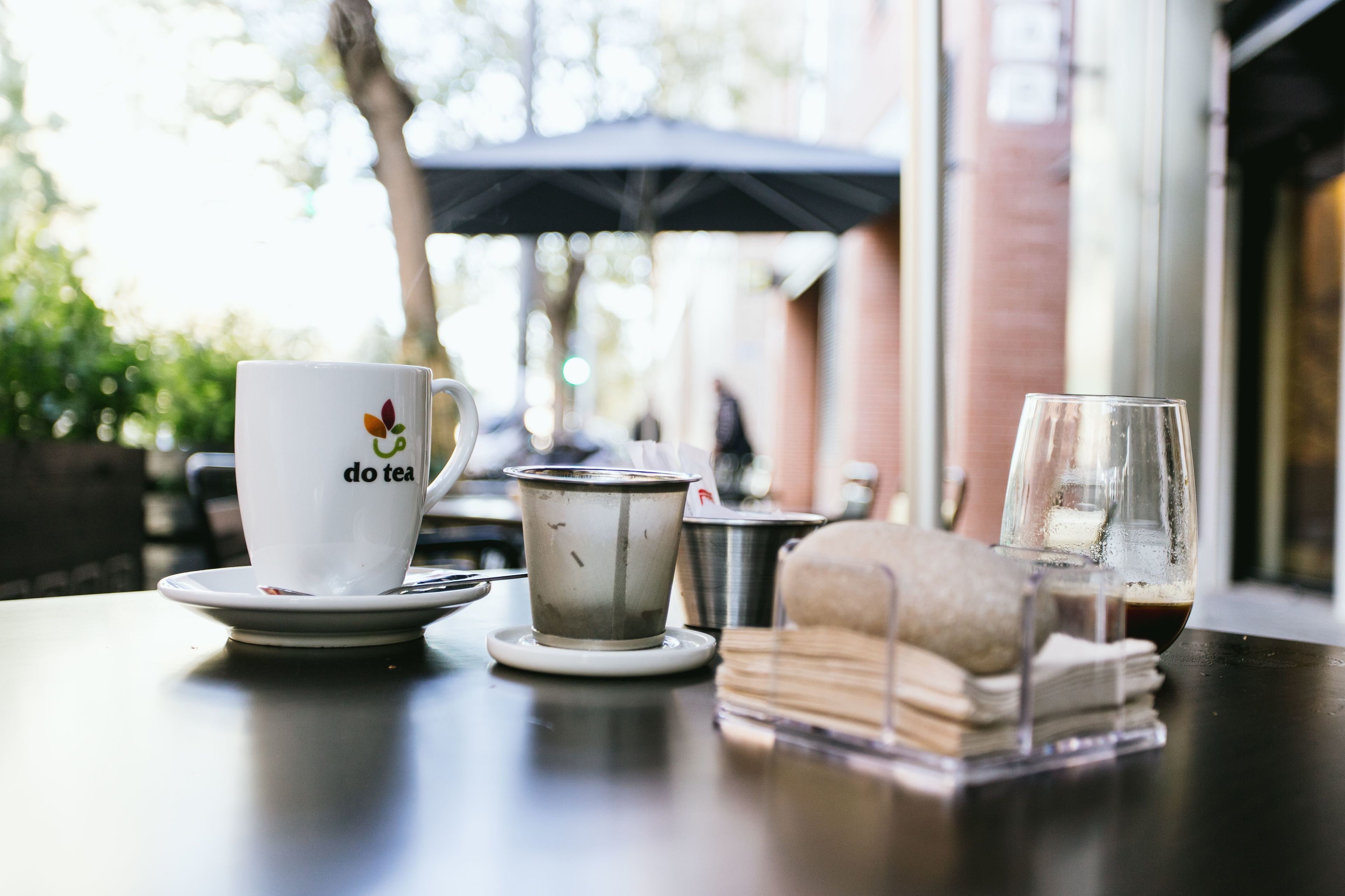 A table with a white cup labeled "do tea," a metal tea pot, a glass with a beverage, and a stack of napkins.