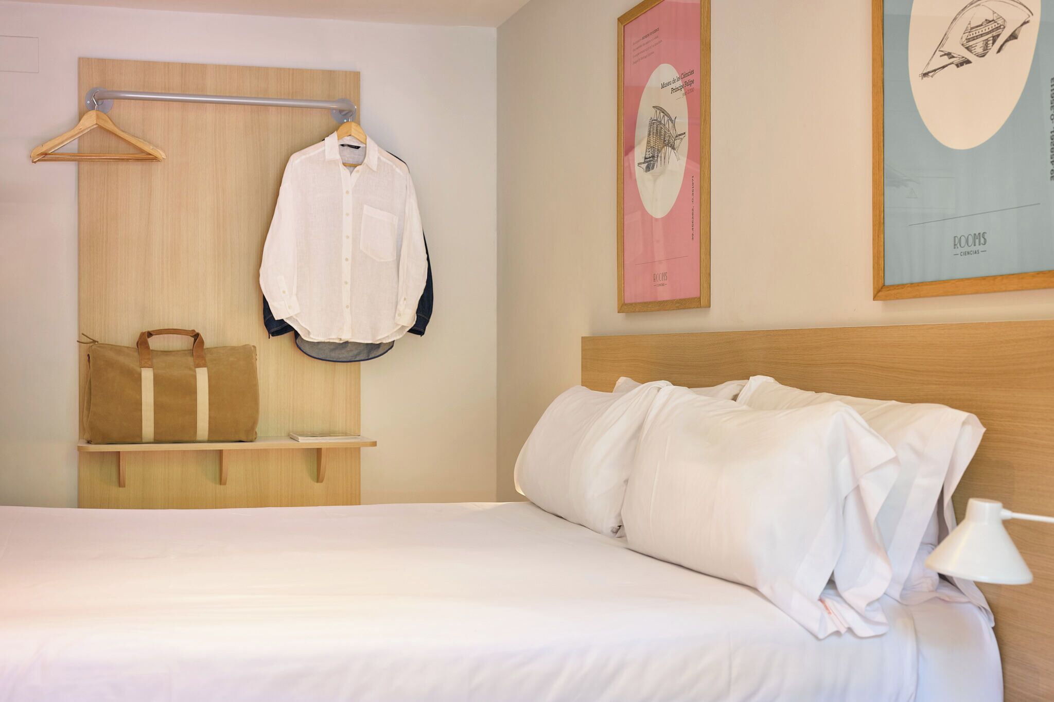 A hotel room with a wooden headboard, a bed with white linens, a hanging clothes rack with a white shirt and a brown bag, and a framed picture on the wall.