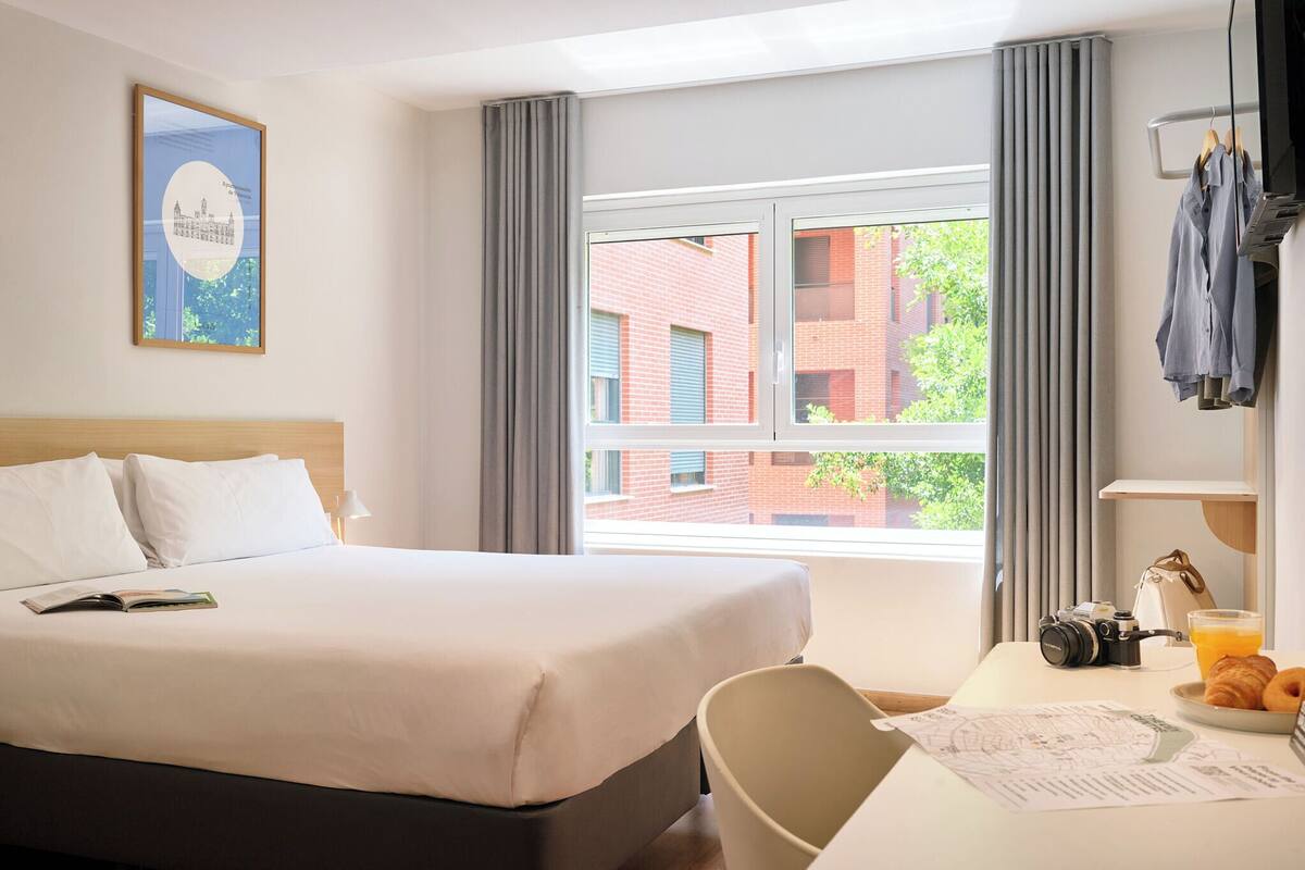 A hotel room with a bed, a desk with a camera, a cup of coffee, and a window with a view of buildings.