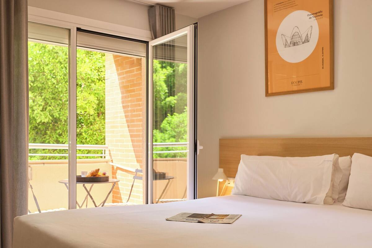 Une chambre d’hôtel avec un lit, une fenêtre donnant sur un paysage verdoyant, une petite table avec une chaise et un tableau encadré au mur.