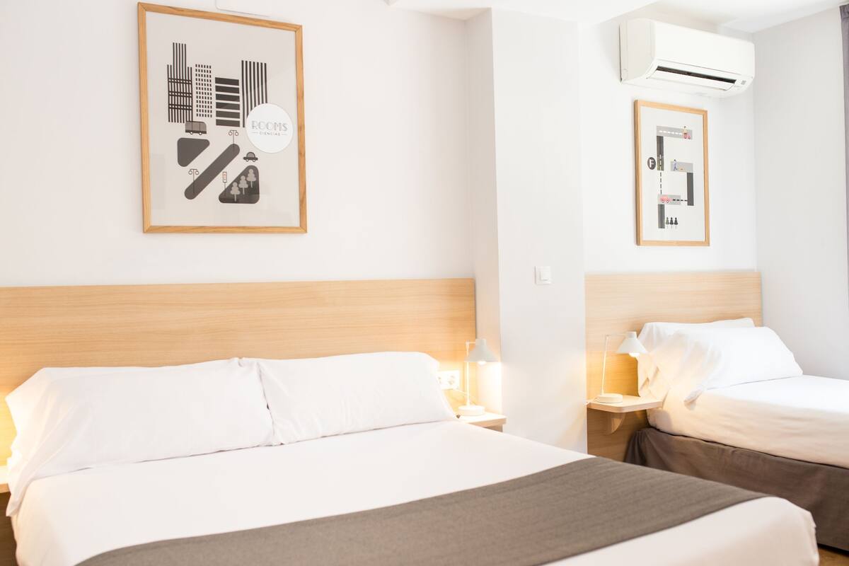 A hotel room with two beds, a framed picture on the wall, an air conditioning unit, and a bedside table with a lamp.