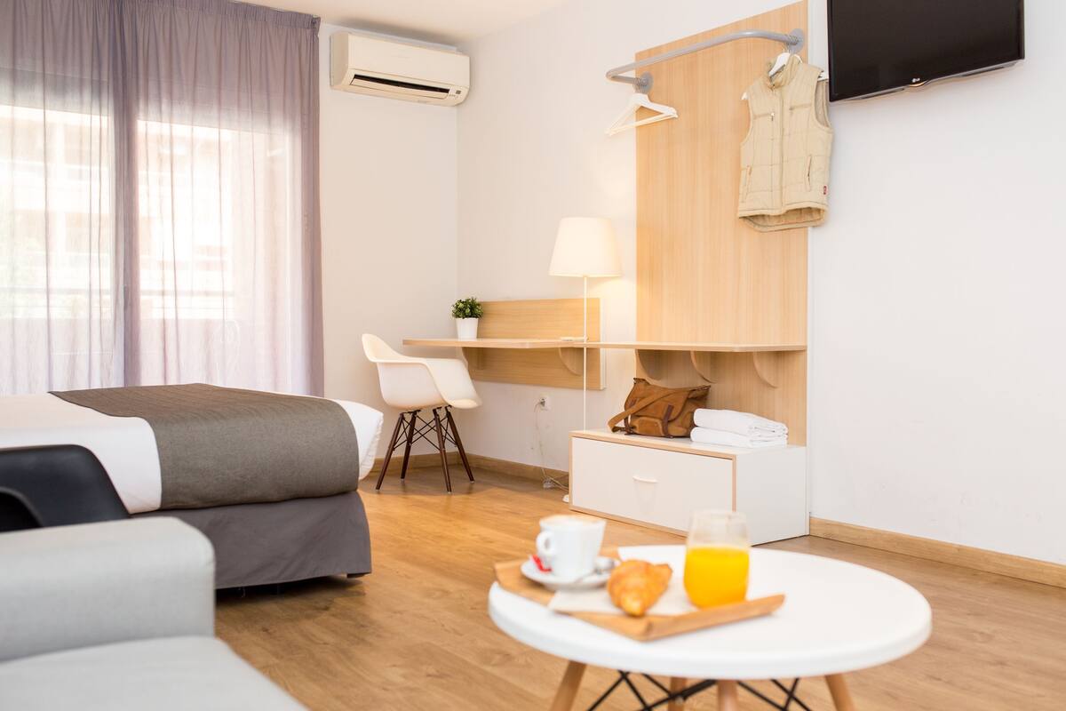 Une chambre d’hôtel moderne équipée d’un lit, d’un bureau, d’une chaise, d’une petite table pour le petit-déjeuner et d’un téléviseur fixé au mur.