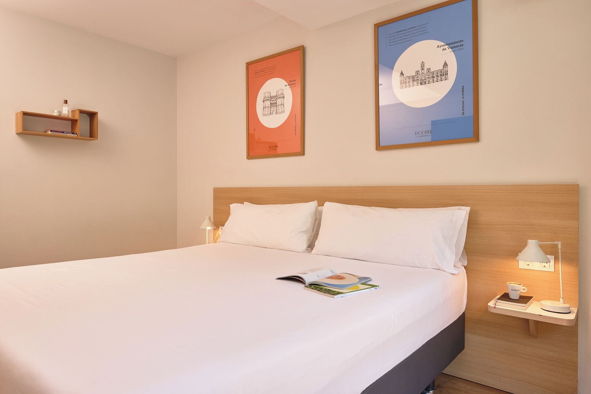 Een net opgemaakt bed met witte lakens, een nachtkastje met een lamp, en twee ingelijste posters aan de muur.