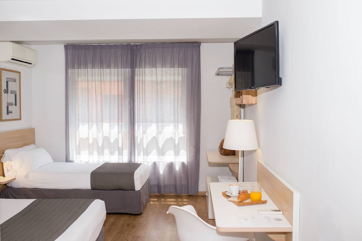 Une chambre d’hôtel avec deux lits, un bureau avec une lampe, une télévision à écran plat et un plateau de petit-déjeuner avec du jus d’orange.