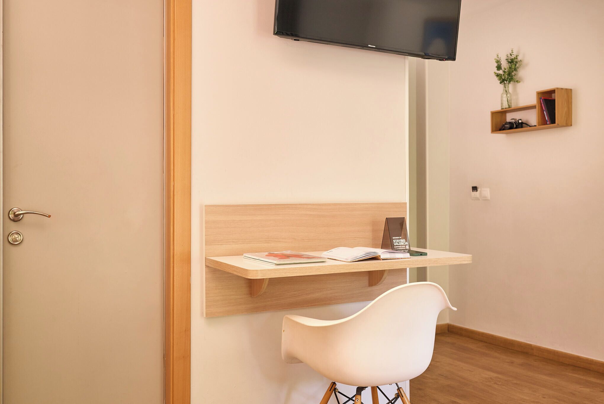 Una habitación moderna con un escritorio de madera, una silla blanca, un televisor montado y un estante pequeño con libros y una planta.