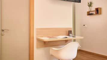 Una habitación moderna con un escritorio de madera, una silla blanca, un televisor montado y un estante pequeño con libros y una planta.
