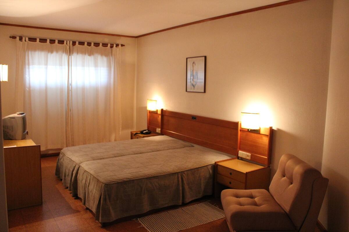 Habitación de hotel con cama doble, lámparas de noche, cabecera de madera, un escritorio pequeño con lámpara, una silla y una ventana con cortinas.