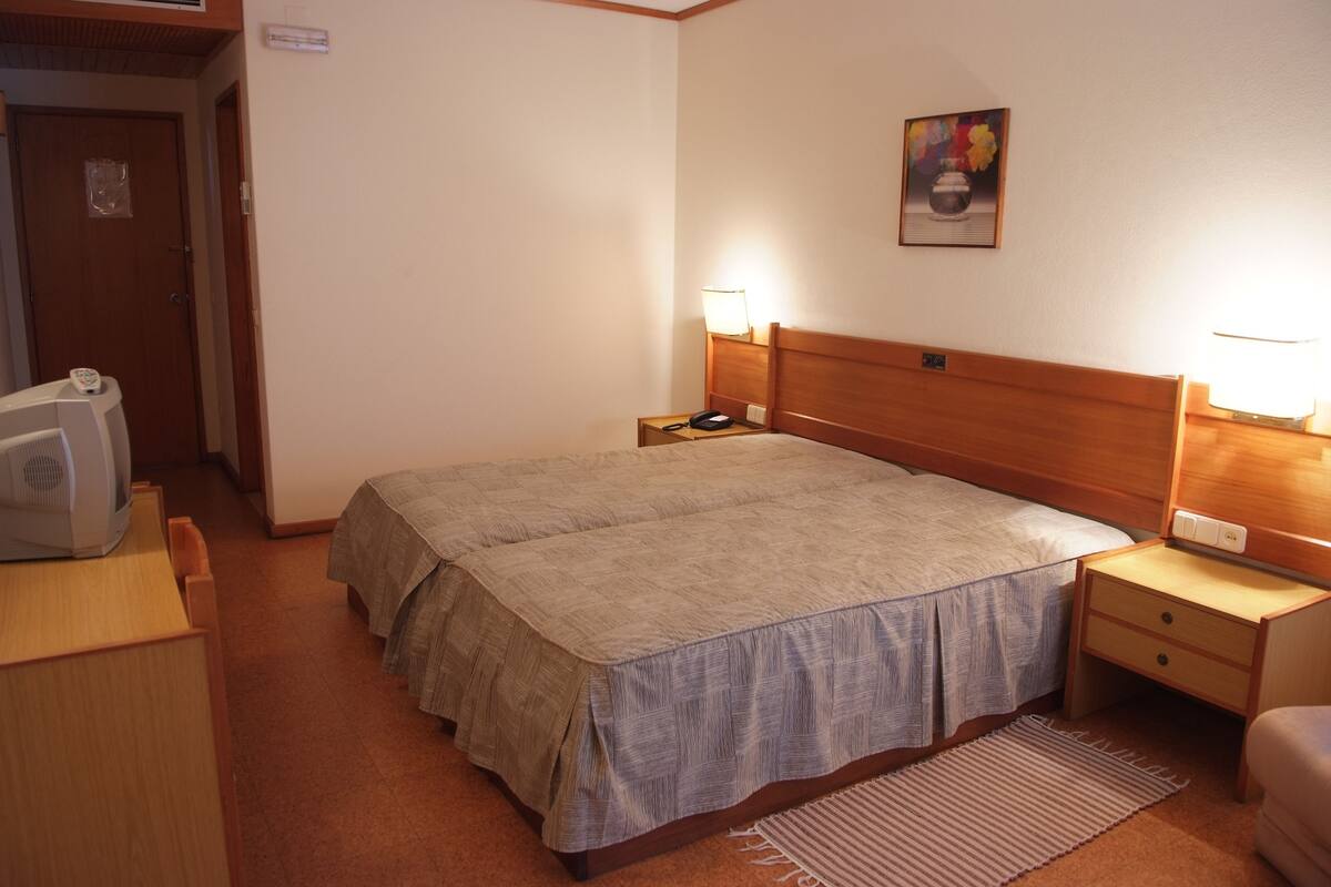 Habitación de hotel con cama doble, lámparas de noche, cabecera de madera, mesita de noche con teléfono y un pequeño escritorio con lámpara, además de un cuadro en la pared.