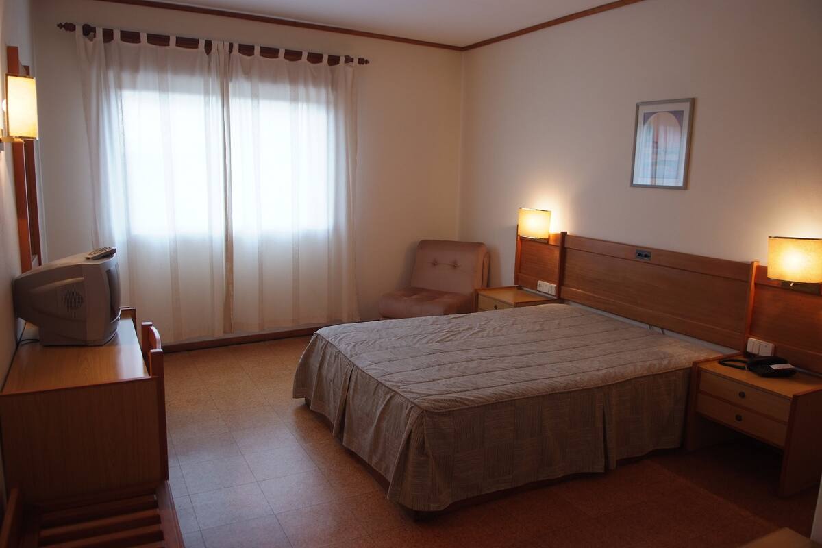 Habitación de hotel con cama, una silla, escritorio con lámpara y un cuadro en la pared.