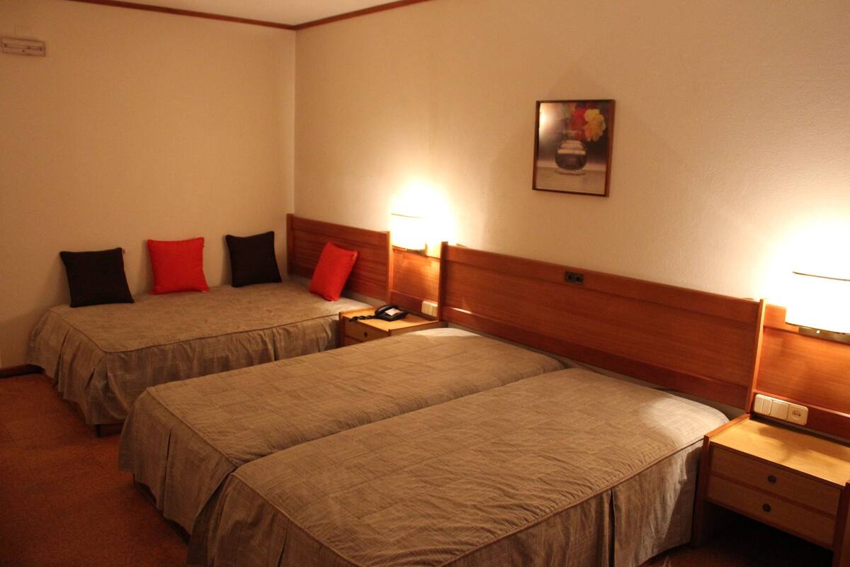Habitación de hotel con dos camas individuales, cada una con cabecero de madera y una mesita de noche. Se observa una mesita con lámpara y teléfono en un lado, y una lámpara de pared sobre las camas.
