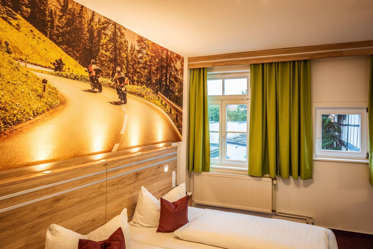 A hotel room with a large mural of a mountain road, a bed with white and red pillows, a window with green curtains, and a view of a parking lot.