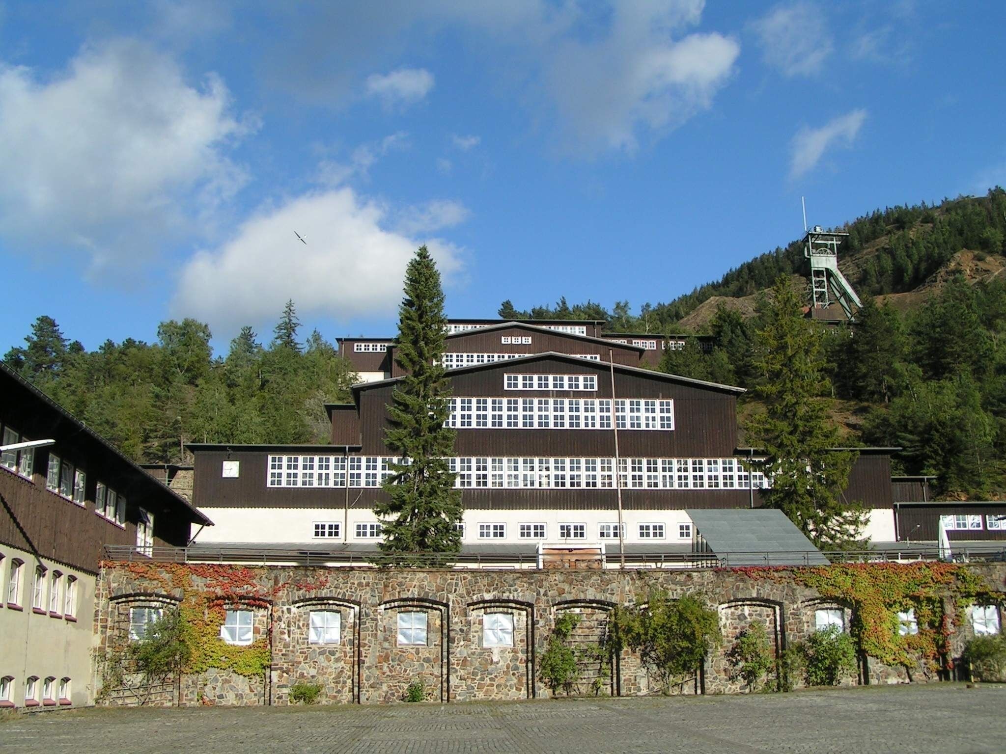 Un grand bâtiment doté de nombreuses fenêtres et d’une façade en pierre, entouré d’arbres et situé dans un paysage vallonné.
