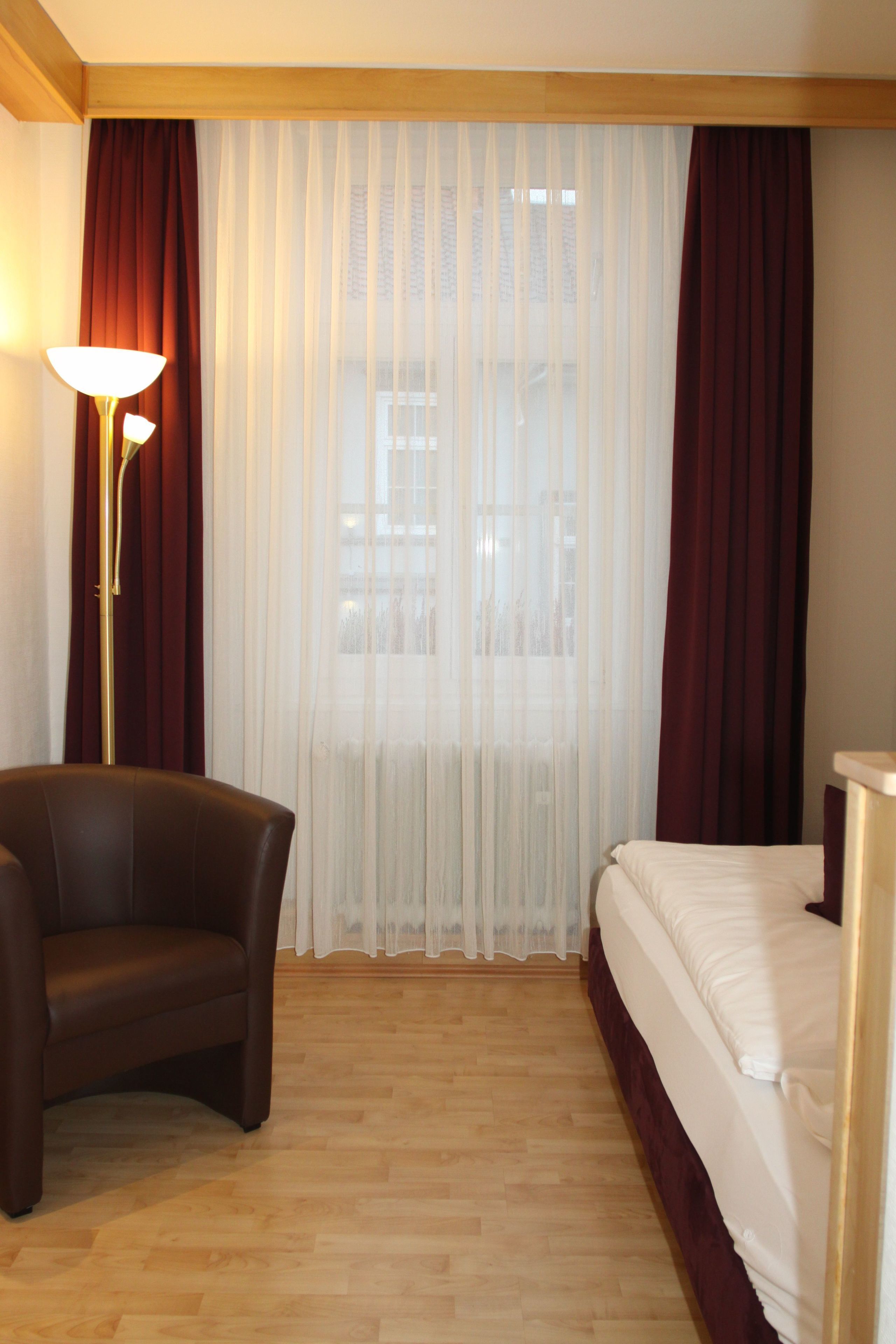 A hotel room with a bed, a chair, a floor lamp, and sheer curtains.