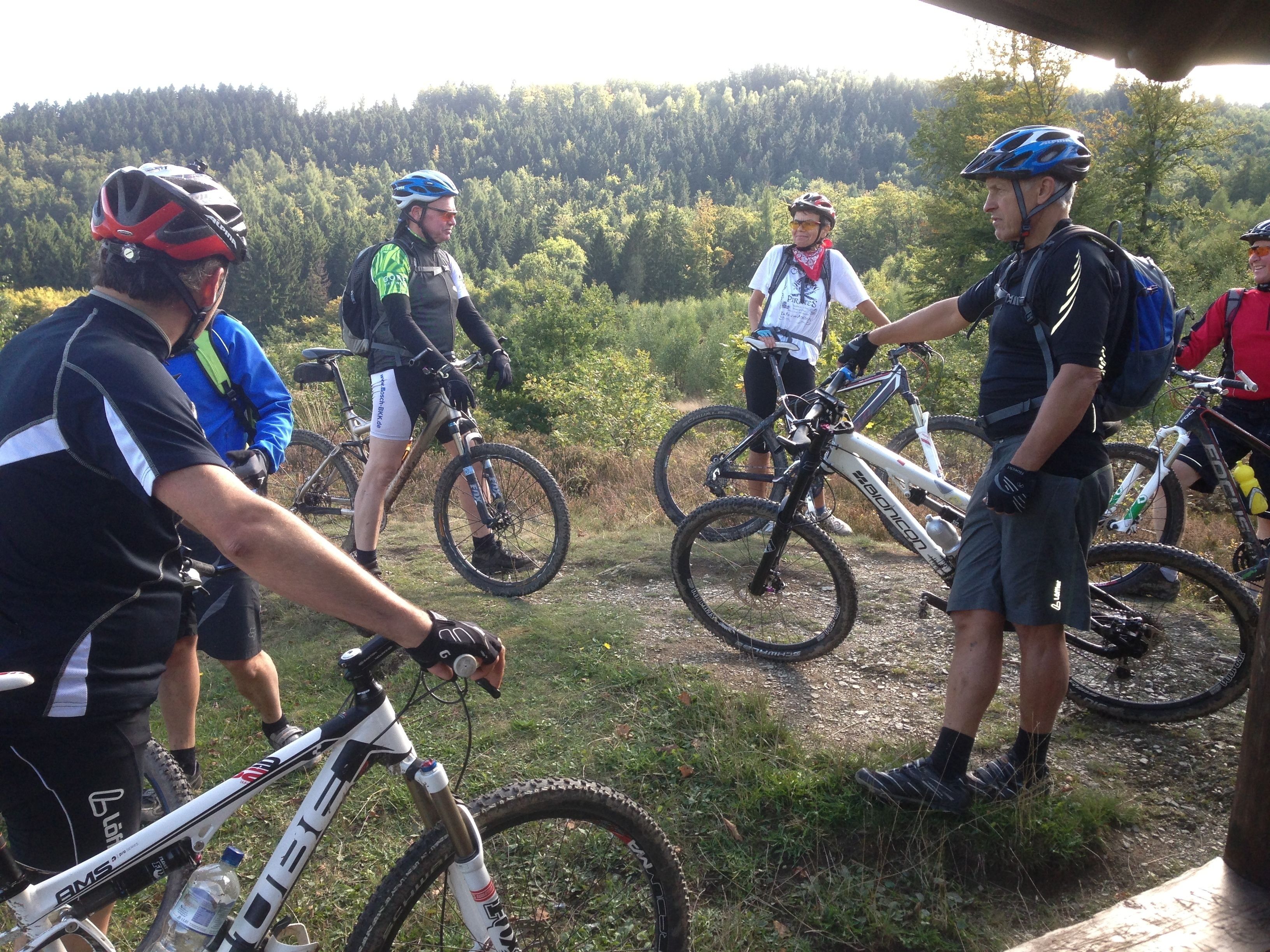 A group of mountain bikers gathered around their bikes in a natural setting with trees in the background.