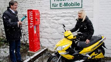 Uma pessoa sentada num scooter amarelo ao lado de uma estação de carregamento vermelha, com uma placa acima que diz 'Reserviert für E-Mobile'.