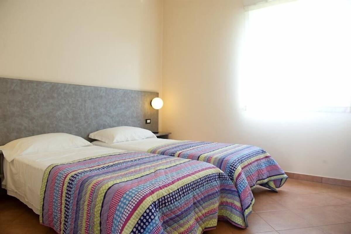 A room with two beds, each with a colorful quilt, a grey headboard, and a wall-mounted light fixture.