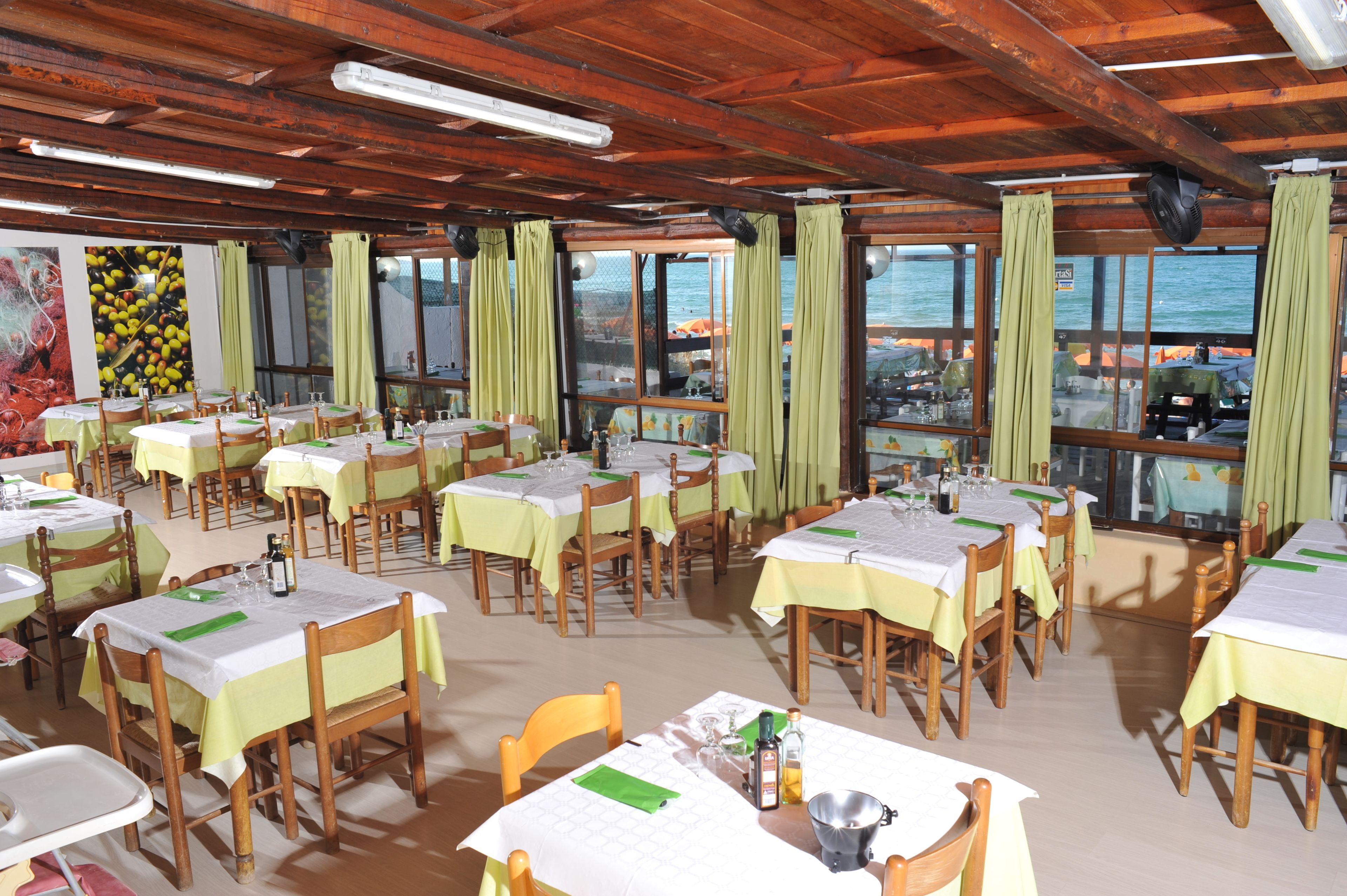 Um restaurante com várias mesas e cadeiras, janelas amplas com cortinas e vista para o mar.