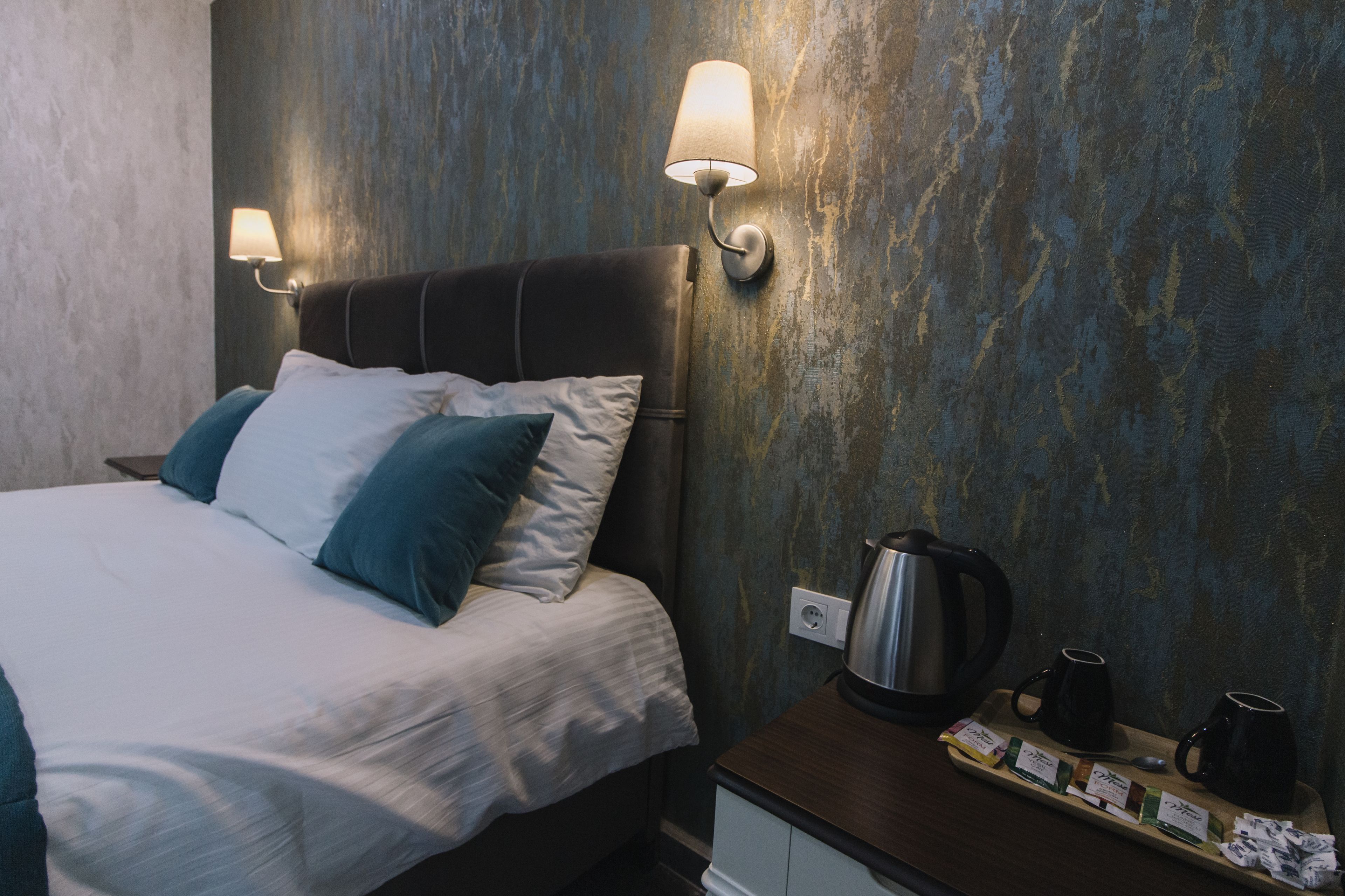 A neatly made bed with pillows, a bedside table with a kettle and mugs, and wall-mounted lamps.