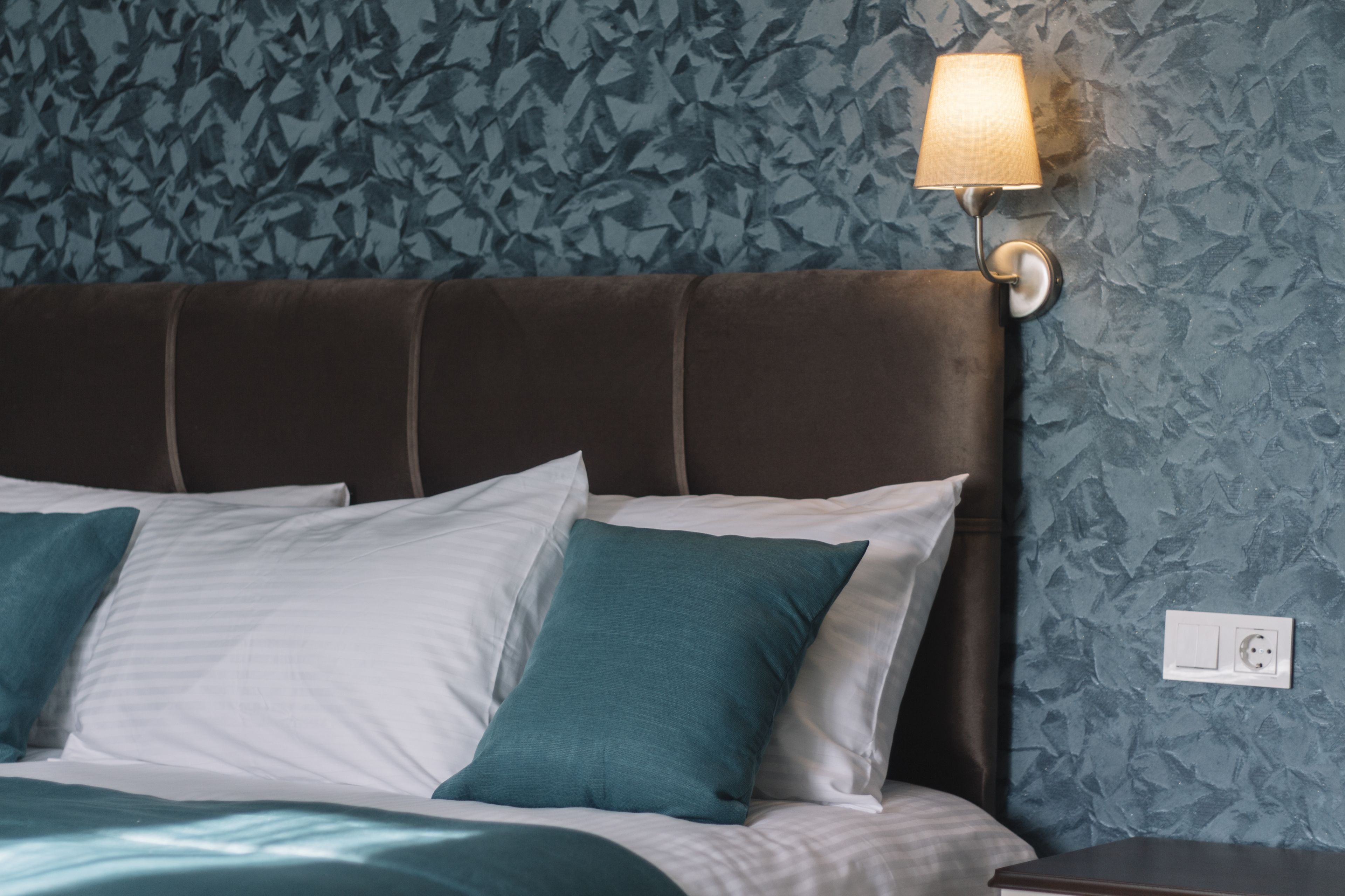 Una cama con almohadas blancas y azul turquesa, un cabecero marrón y una pared con textura.