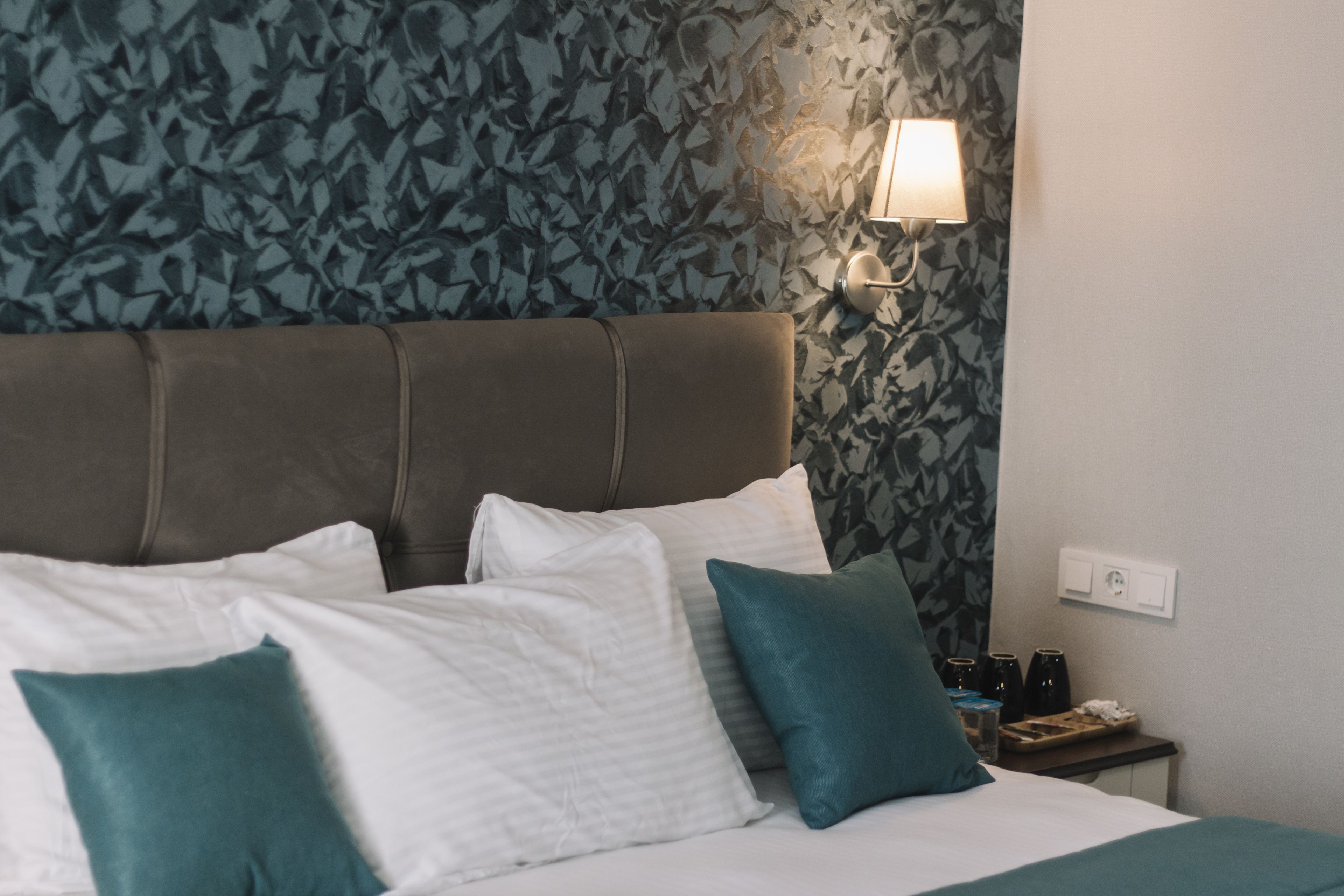 A neatly made bed with white and teal pillows, a brown headboard, and a wall with a leafy pattern.