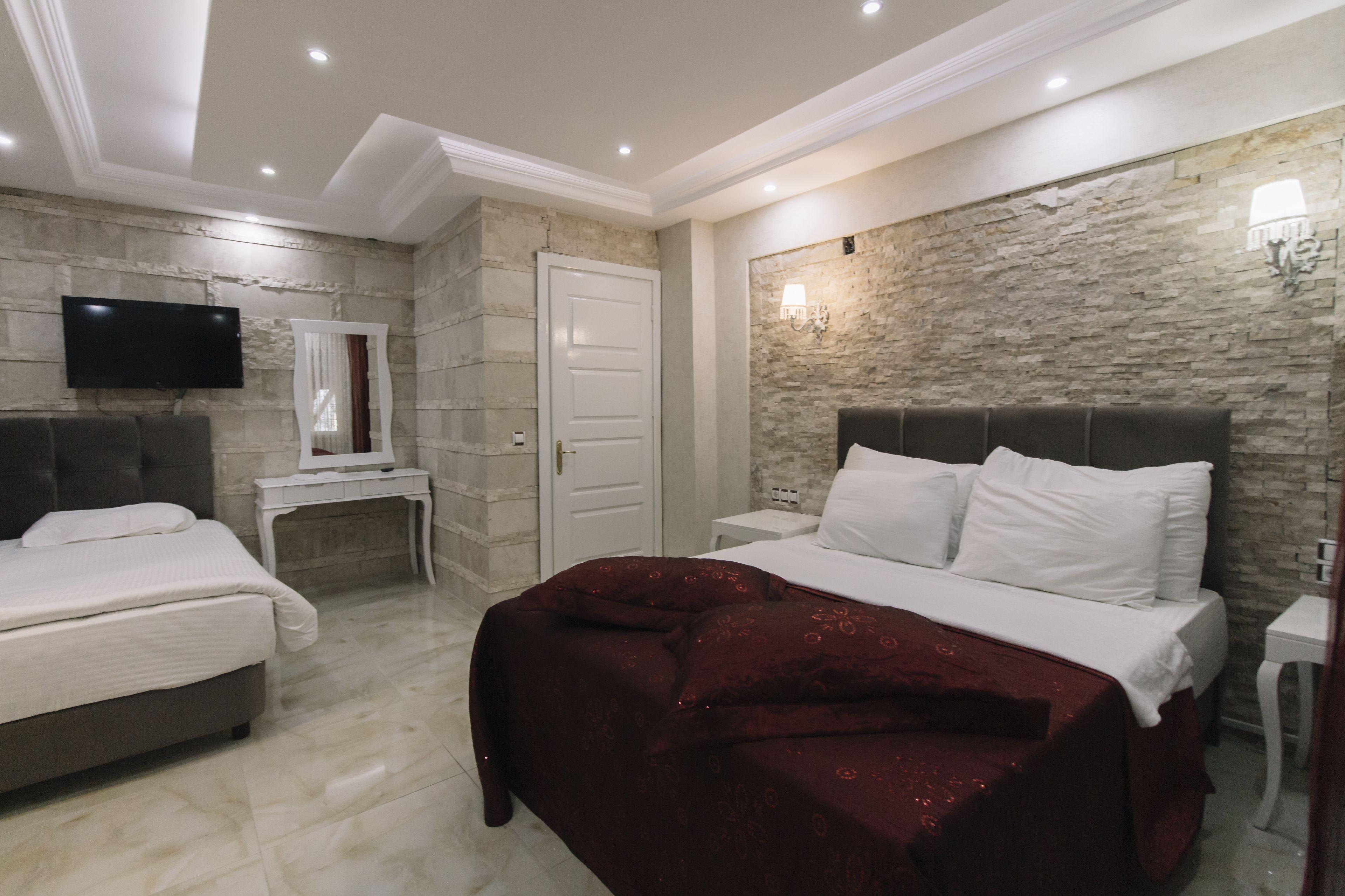 A hotel room with a marble floor, a stone accent wall, a flat-screen TV mounted on the wall, a bed with white bedding and a red throw, and a bedside table with a lamp.