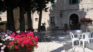 An outdoor seating area with tables and chairs, surrounded by stone buildings with arched windows and green shutters. - Residenza del Marchese (Gualdo Cattaneo)
