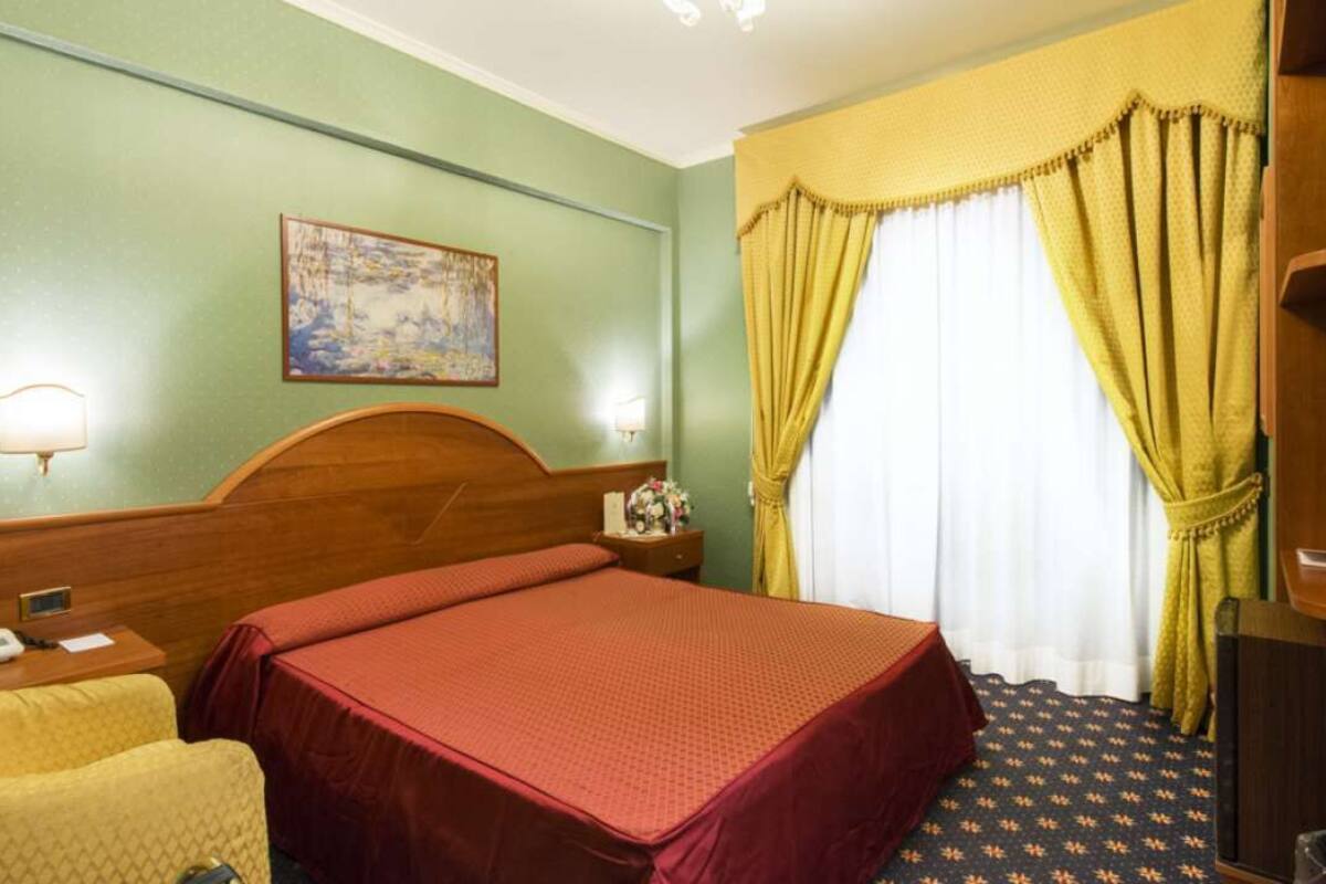 A hotel room with a single bed, a bedside table with a lamp, a chair, and a painting on the wall.