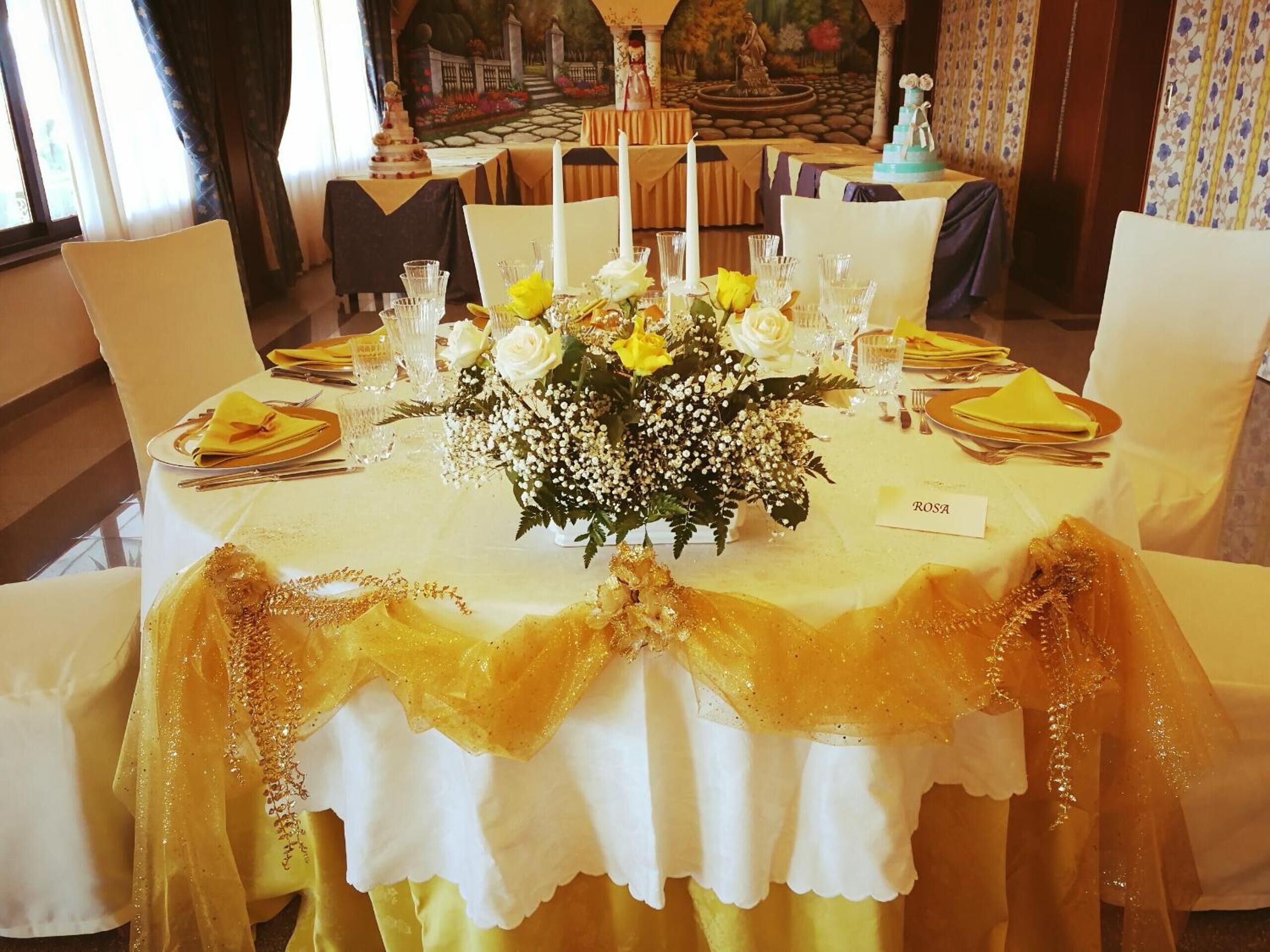 A formal dining table set with white and yellow tablecloths, a centerpiece of yellow roses, and a name card reading "ROSA".