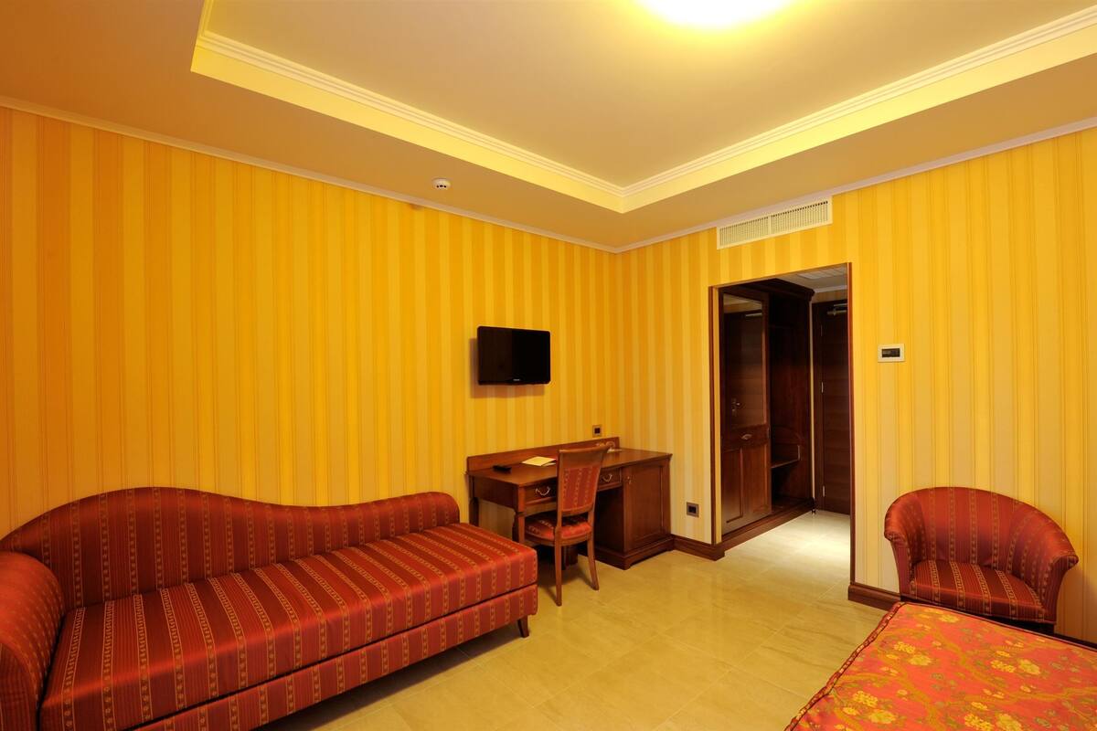 A hotel room with striped yellow walls, a sofa, a desk with a chair, and a television.