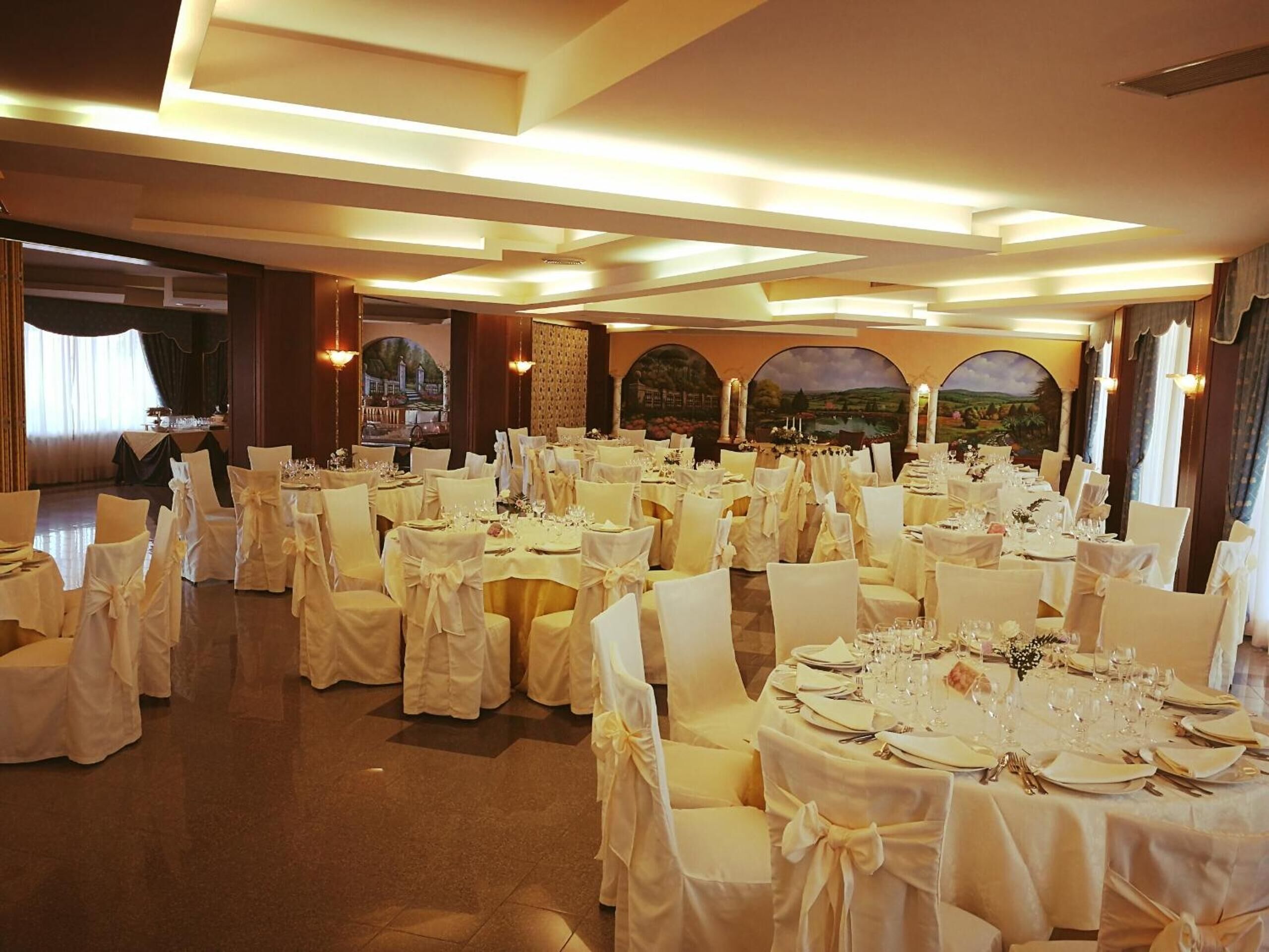Un salón de banquetes con mesas redondas dispuestas para cenar, sillas blancas y vistas a un paisaje pintoresco a través de ventanales arqueados.