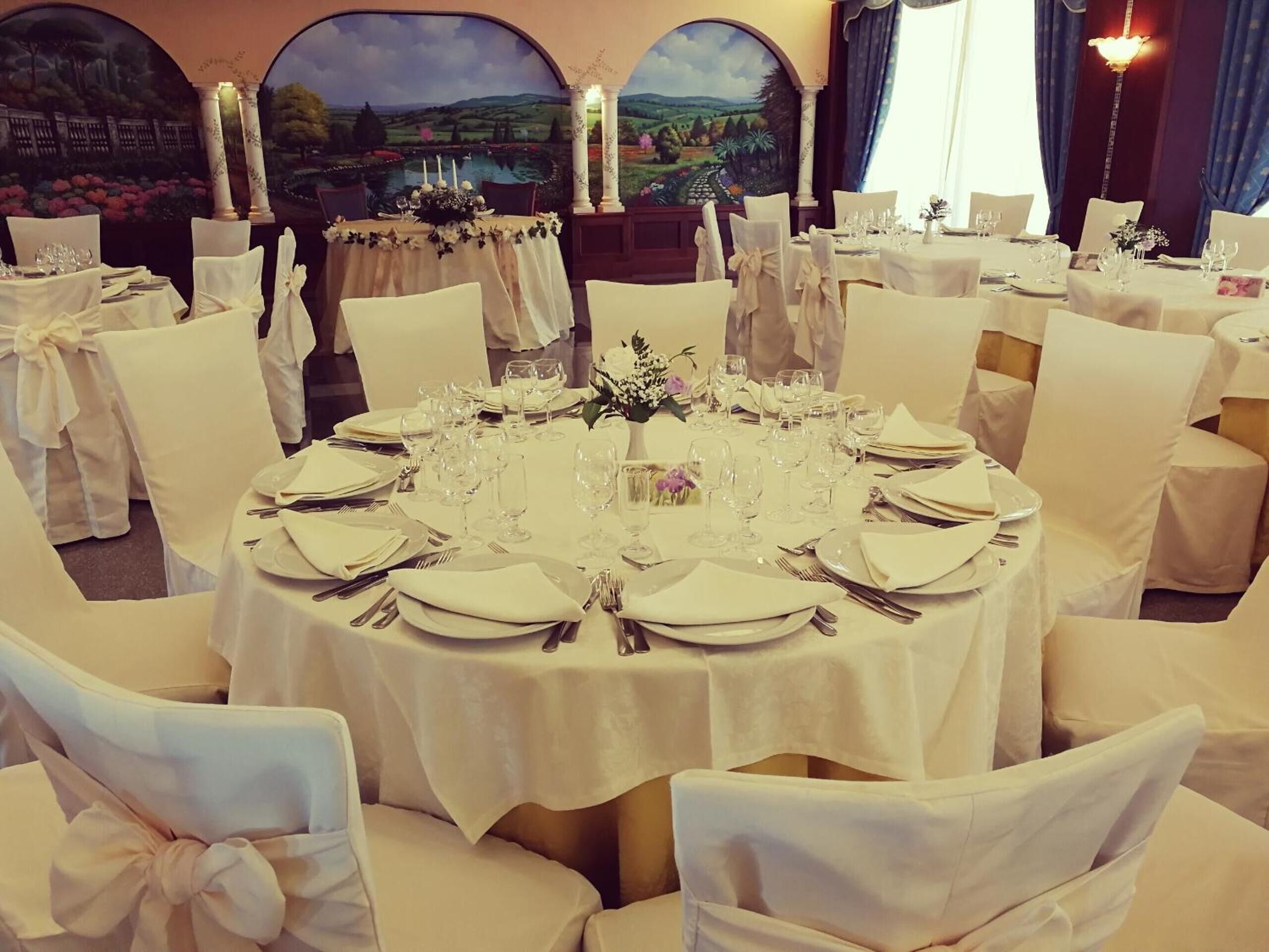 Uma sala de jantar formal com mesas redondas preparadas para refeição, cadeiras brancas com laços e um mural paisagístico ao fundo.