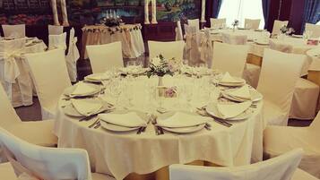 Uma sala de jantar formal com mesas redondas preparadas para refeição, cadeiras brancas com laços e um mural paisagístico ao fundo.