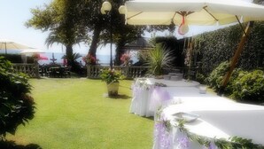Un jardín con mesas y sillas blancas, rodeado de follaje y vista al mar.