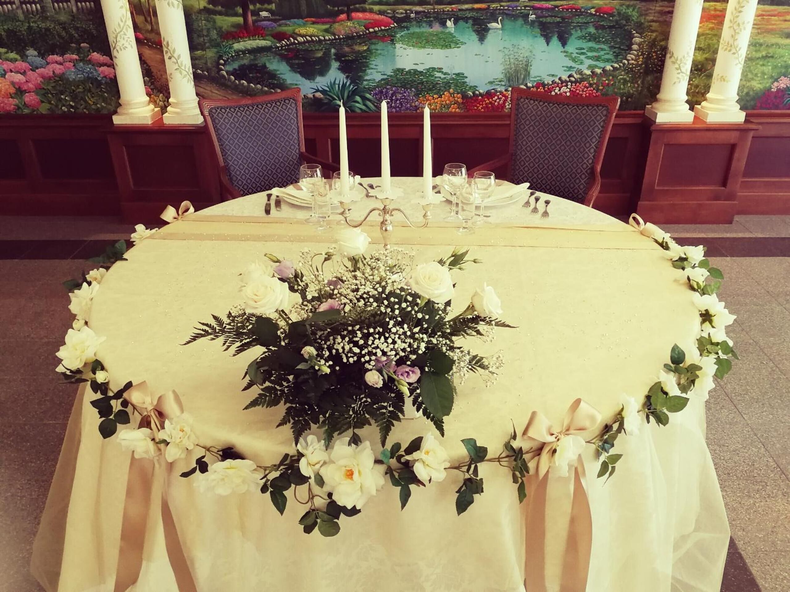 A round dining table with a floral centerpiece, white candles, and a view of a garden through a window.