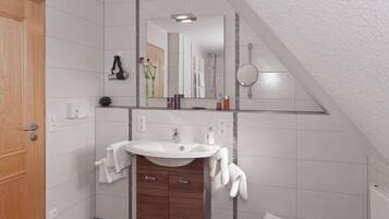 A bathroom with a wooden door, white tiled walls, a white sink with a mirror above it, and a small trash bin.
