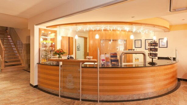 A reception area with a curved wooden counter, a staircase to the left, and a wall with framed certificates. - Hotel Garni Altes Brauhaus (Rothenburg ob der Tauber)