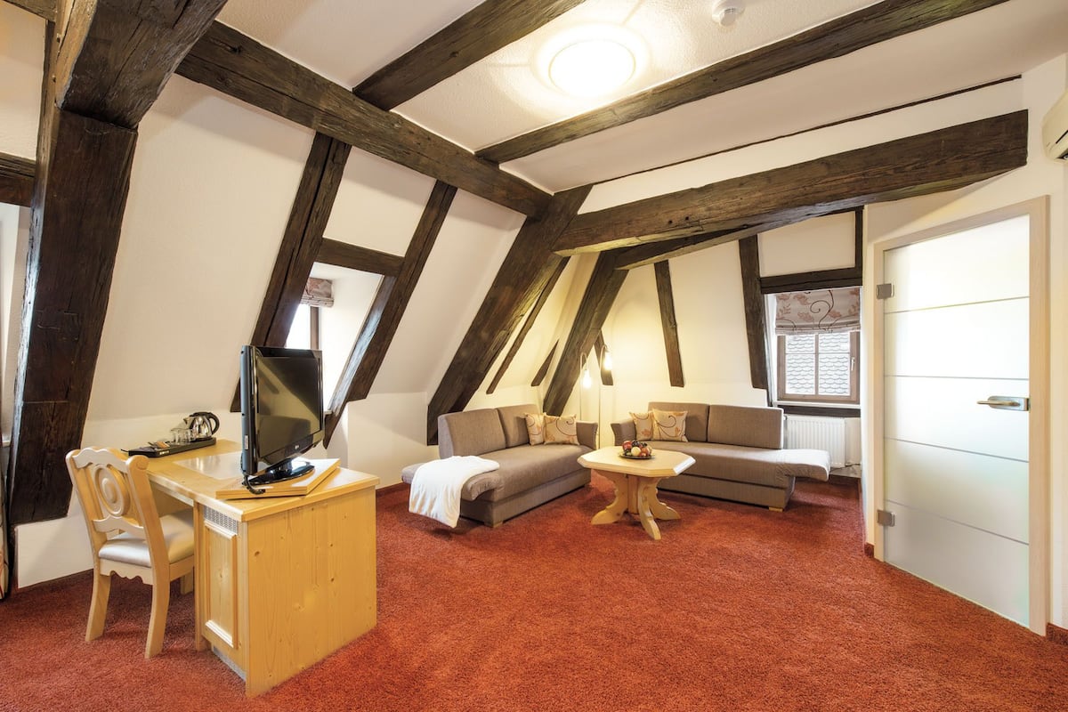Acogedor dormitorio en ático con vigas de madera a la vista, un escritorio de madera con computadora, un sofá y una pequeña mesa redonda con lámpara.