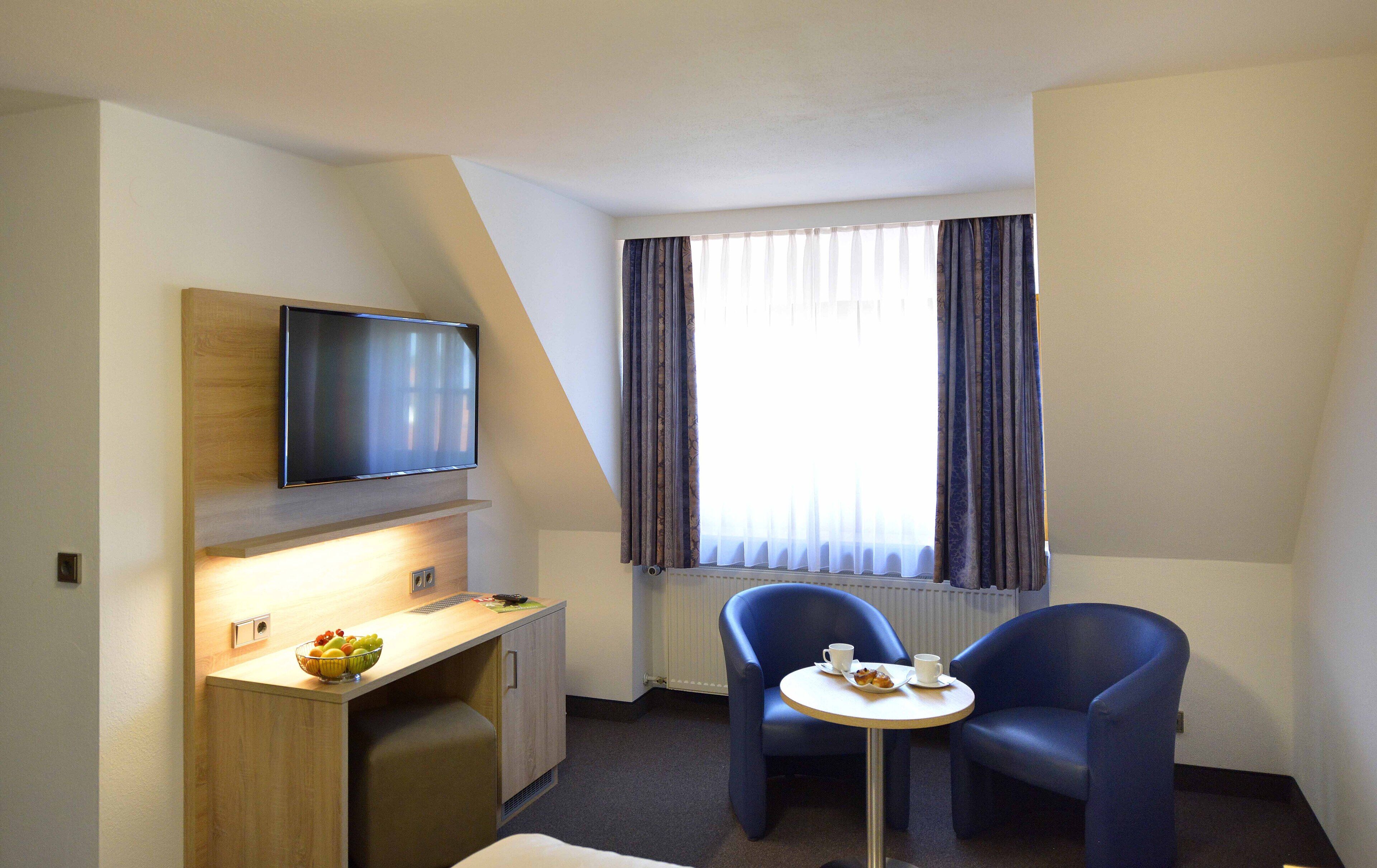 Hotelkamer met een flatscreen televisie aan de muur, een klein houten bureau met een schaal fruit, twee blauwe fauteuils en een ronde tafel met bekers en gebak.