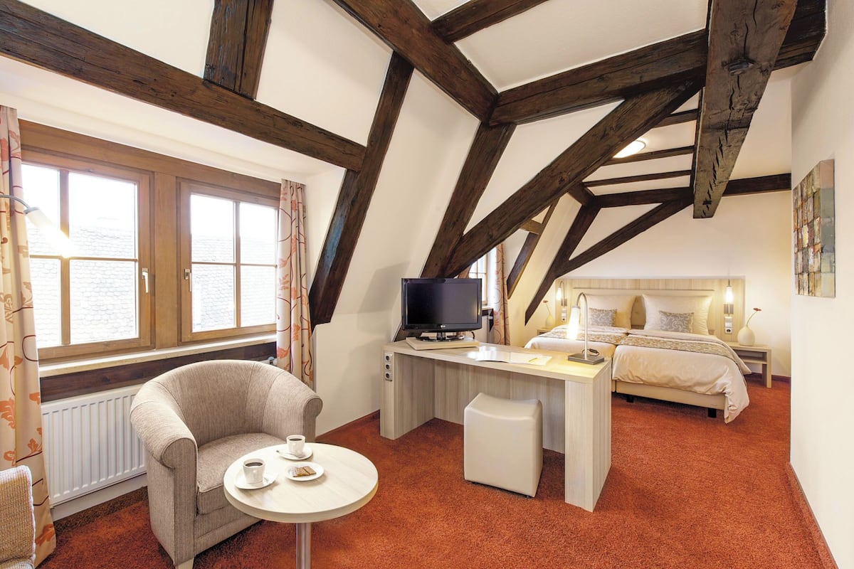 Una habitación con techo alto, vigas de madera a la vista, una cama, un escritorio con computadora, una silla y una ventana con cortinas.