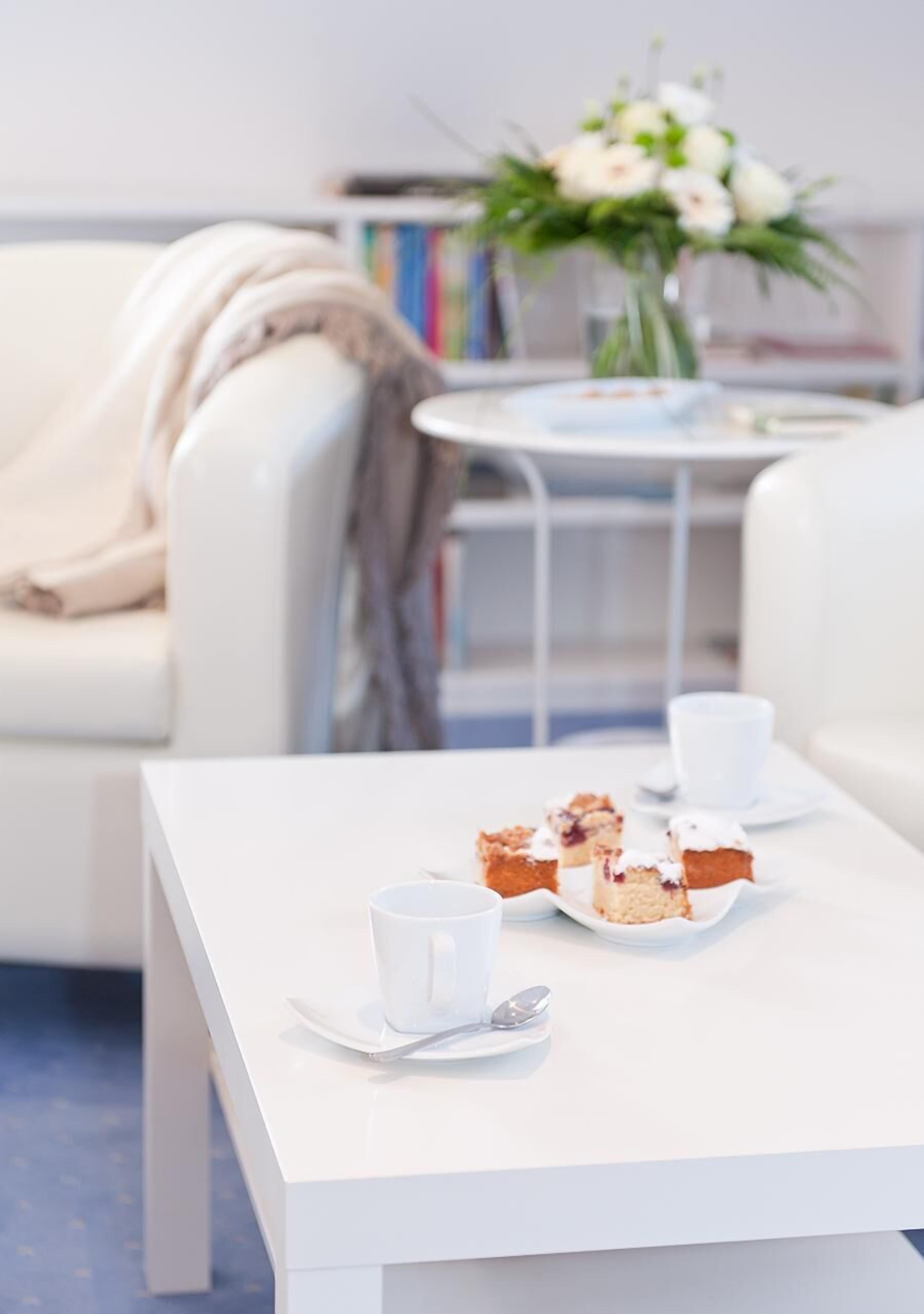 Una mesa blanca con una taza blanca y un plato de comida, un sofá blanco y un sillón blanco en una habitación con una estantería y un jarrón de flores.