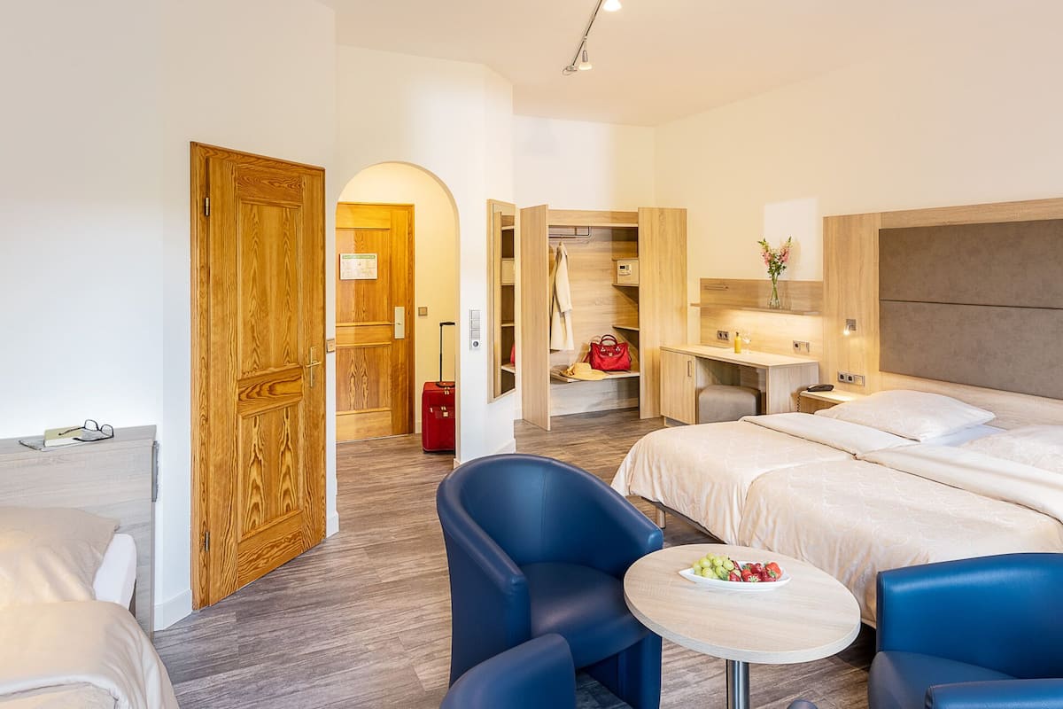 Una habitación de hotel con una cama, dos sillones azules, una mesita redonda con fruta, un escritorio con lámpara y un armario.