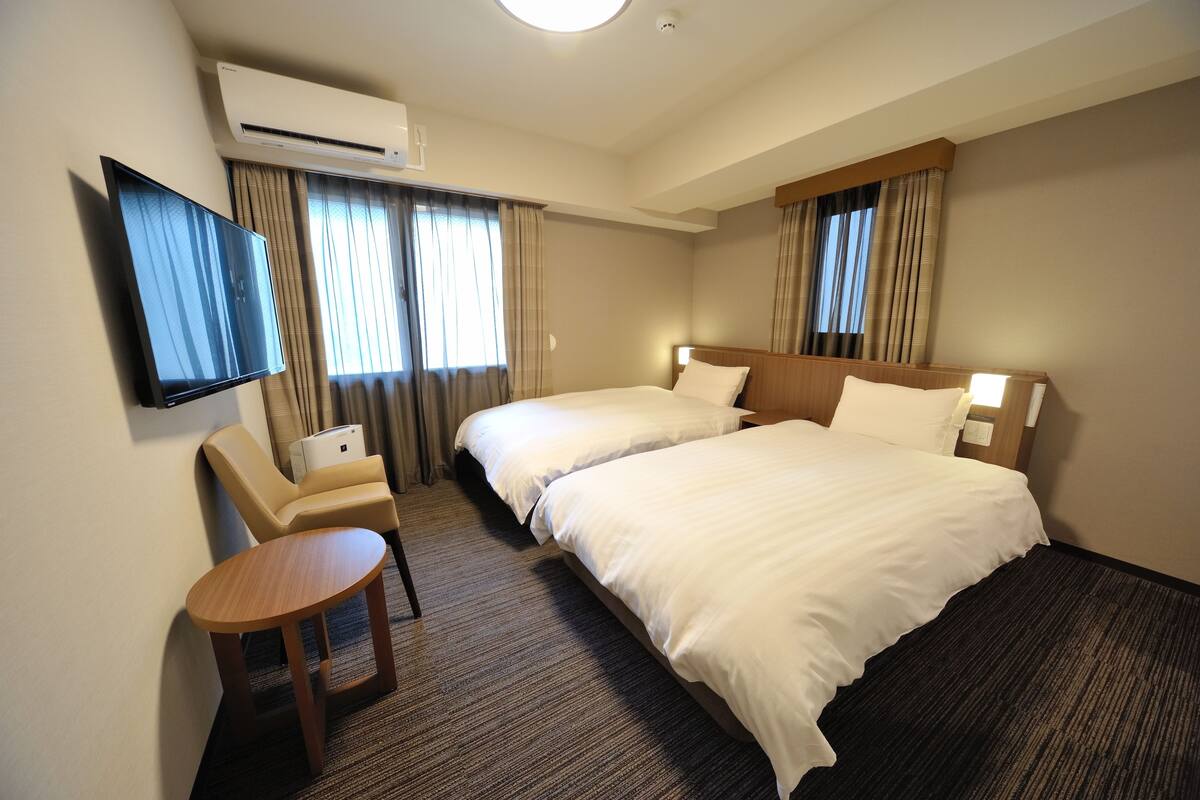 A hotel room with two beds, a TV, a desk, and an air conditioning unit.