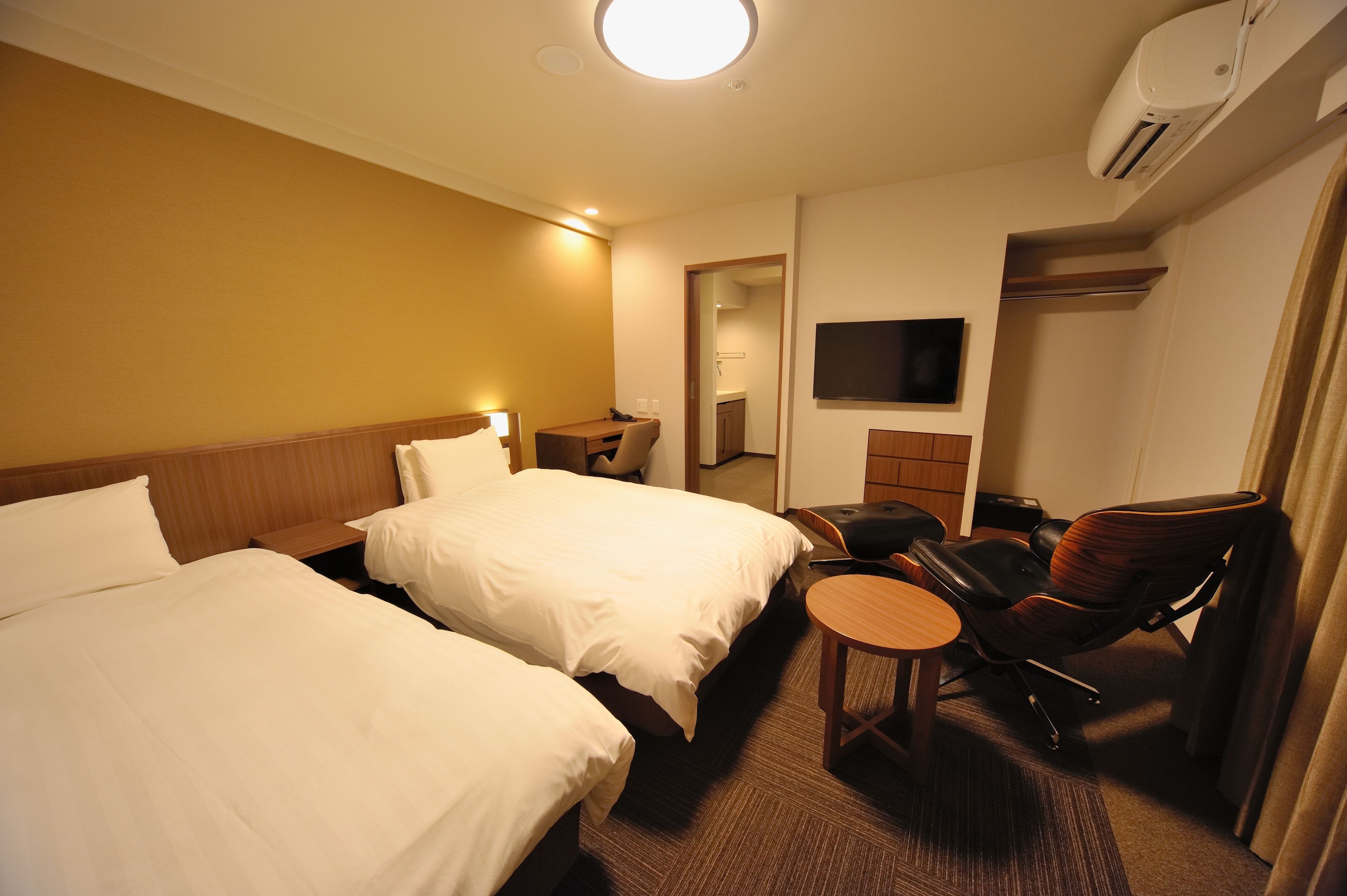 A hotel room with two beds, a desk, a chair, a TV, and an air conditioning unit.