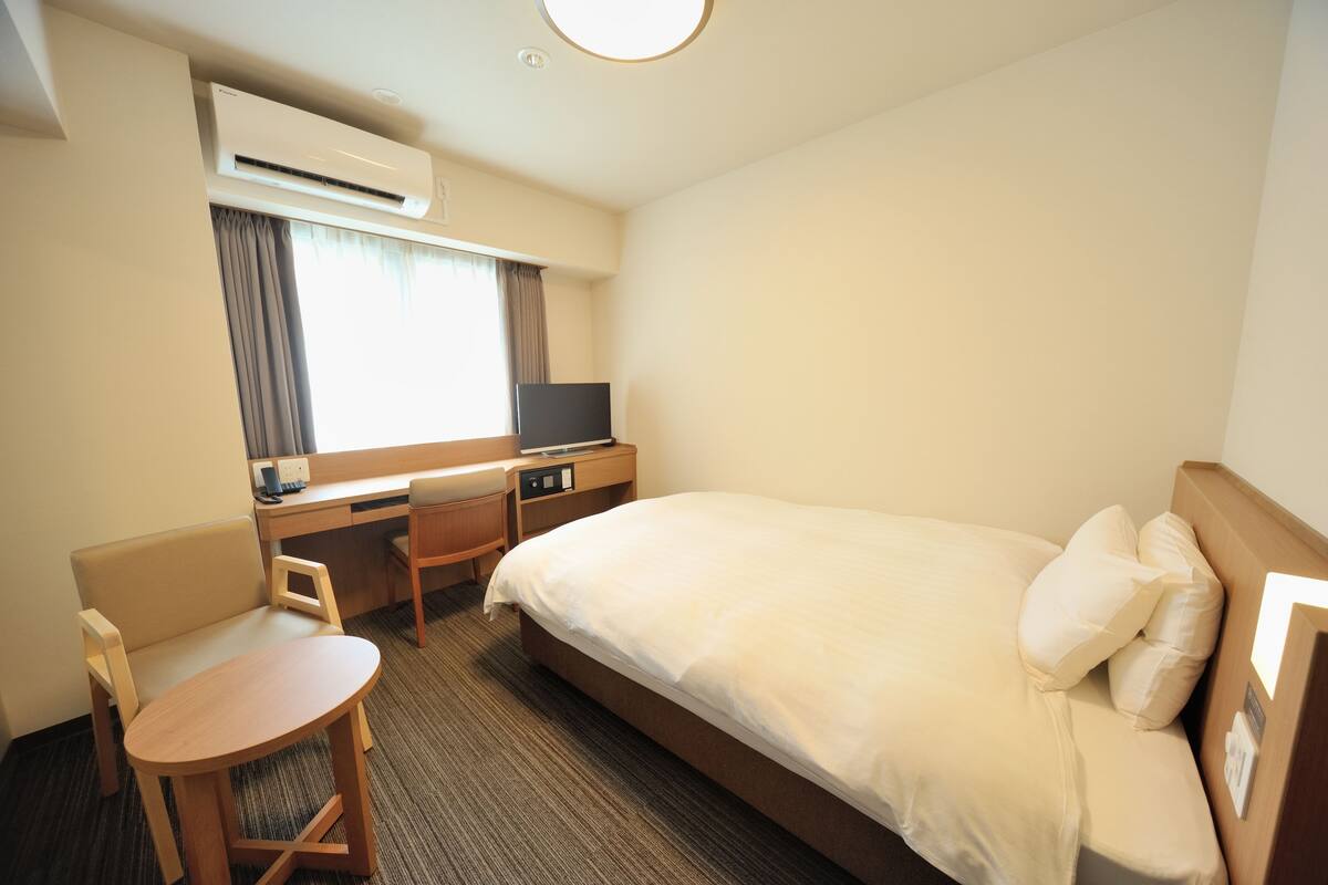 A hotel room with a bed, a desk, a chair, a television, and an air conditioning unit.