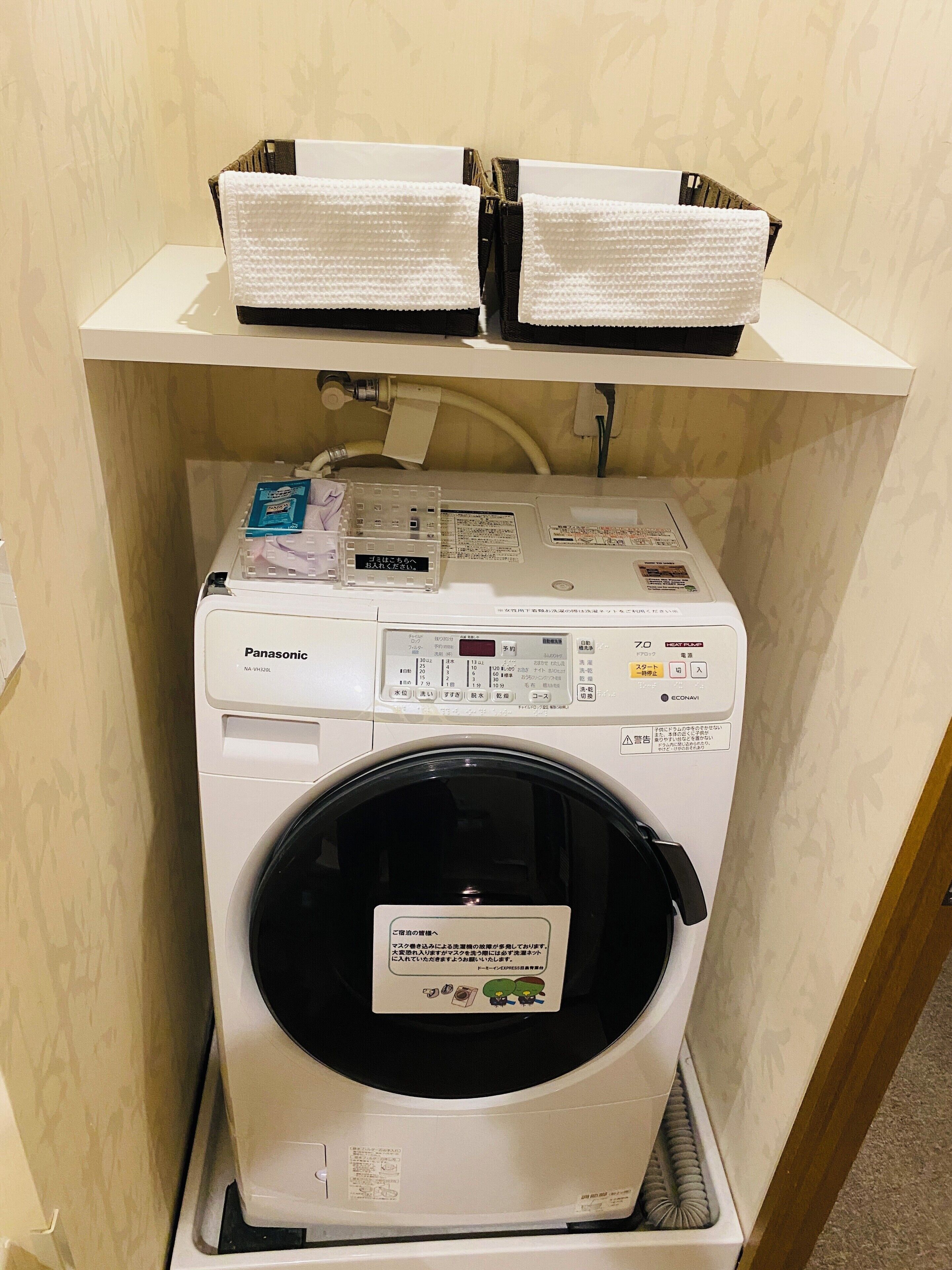 A compact washing machine with a control panel, a small shelf above it, and a power cord plugged in.