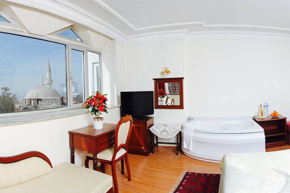A hotel room with a large window, a flat-screen TV, a white bathtub, a wooden cabinet, and a bed with a red pillow.